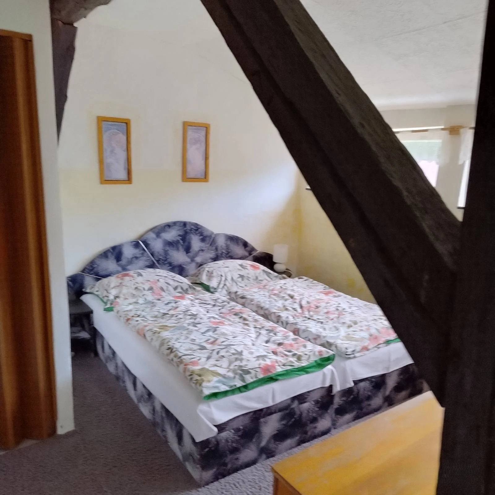 A bedroom with two single beds pushed together, covered with floral blankets, a headboard with a marbled pattern, and a small nightstand with a lamp. Two framed pictures hang on the yellow and white wall, and a window with curtains is visible to the 