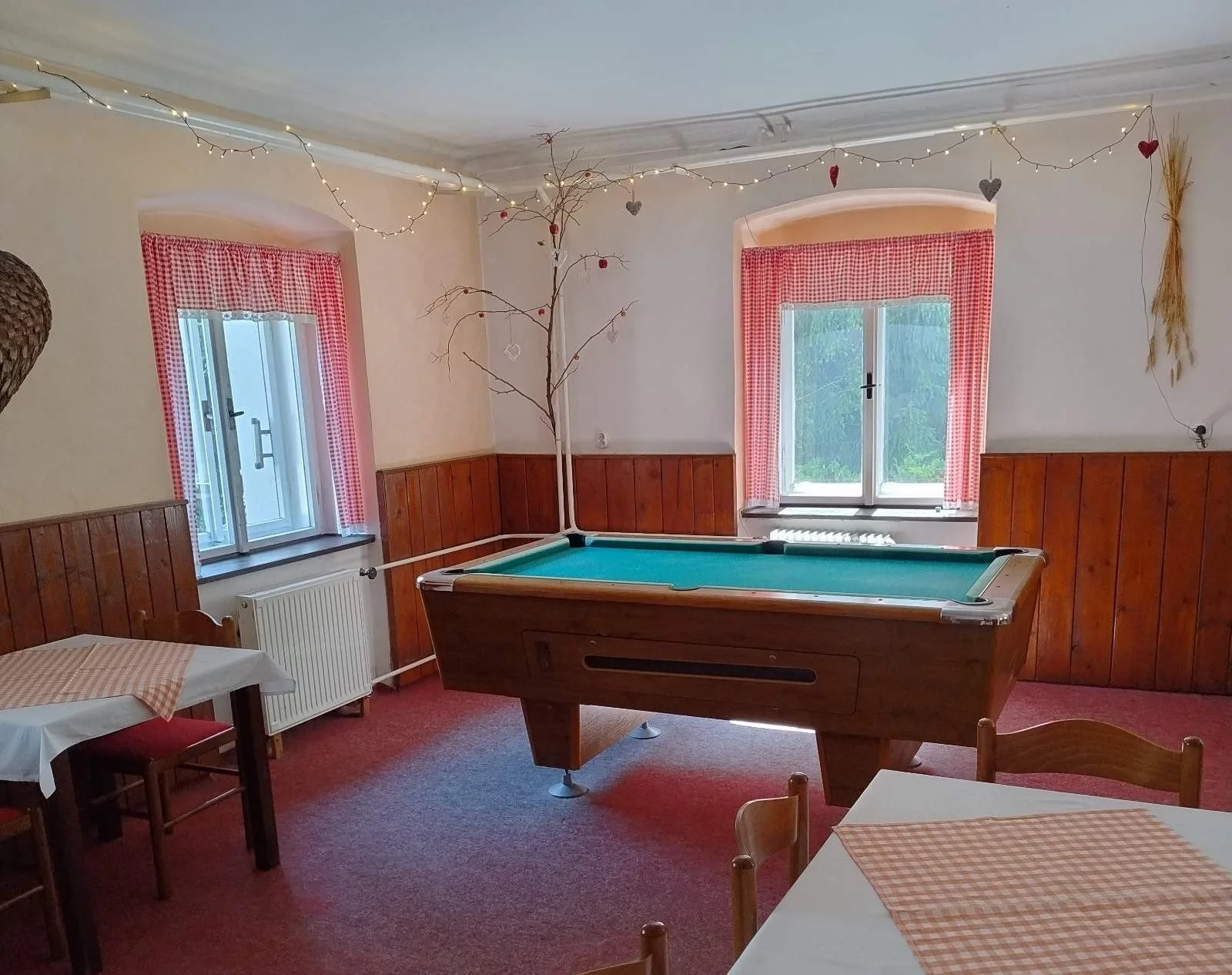 A cozy room with two windows with red checkered curtains, a pool table in the center, and dining tables with checkered tablecloths. The room is decorated with string lights and hanging hearts.