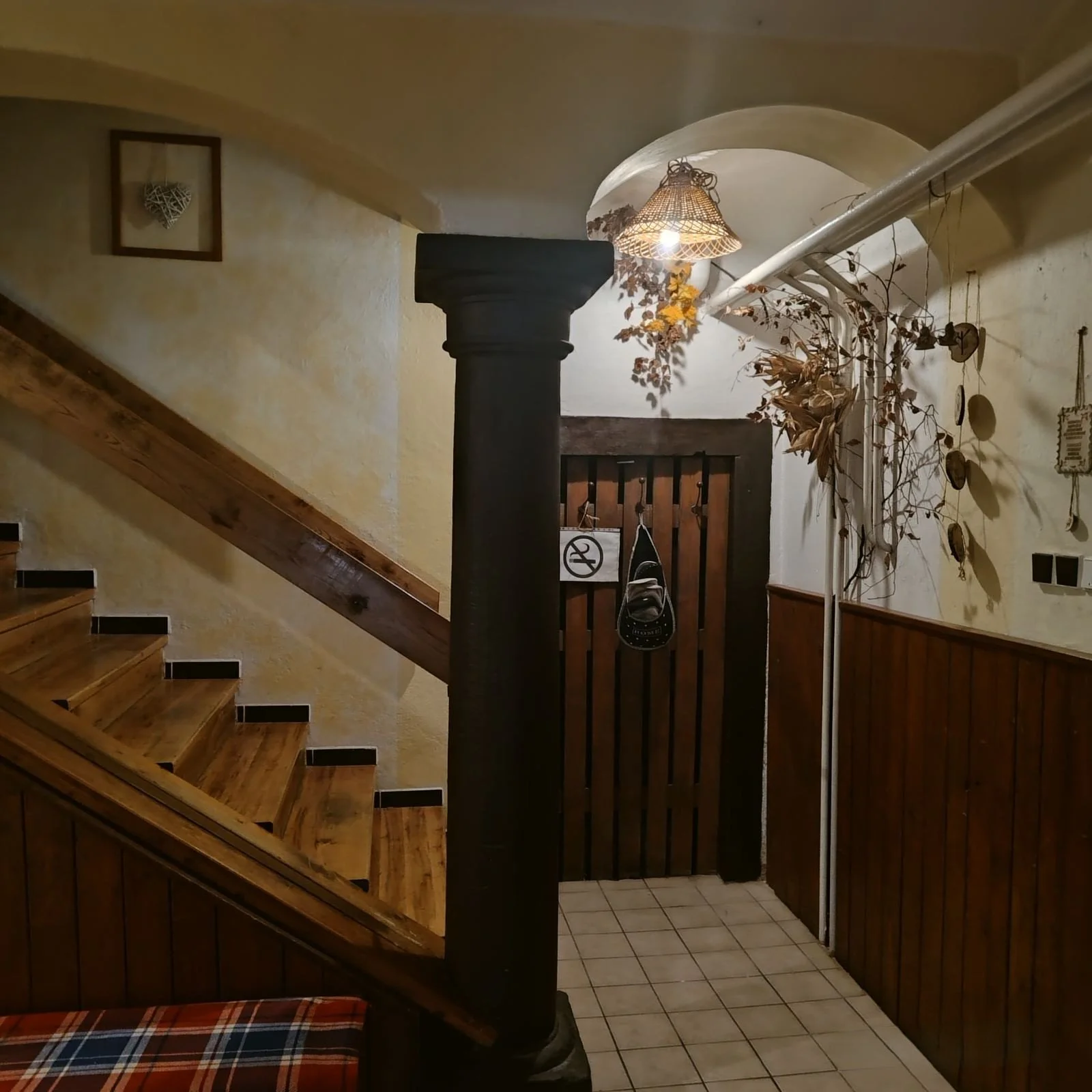 Indoor hallway with wooden staircase, a dark colored column, a framed heart-shaped decoration, dried plants hanging, a coat rack with bags, and a door with a 'no smoking' sign, illuminated by a ceiling light.
