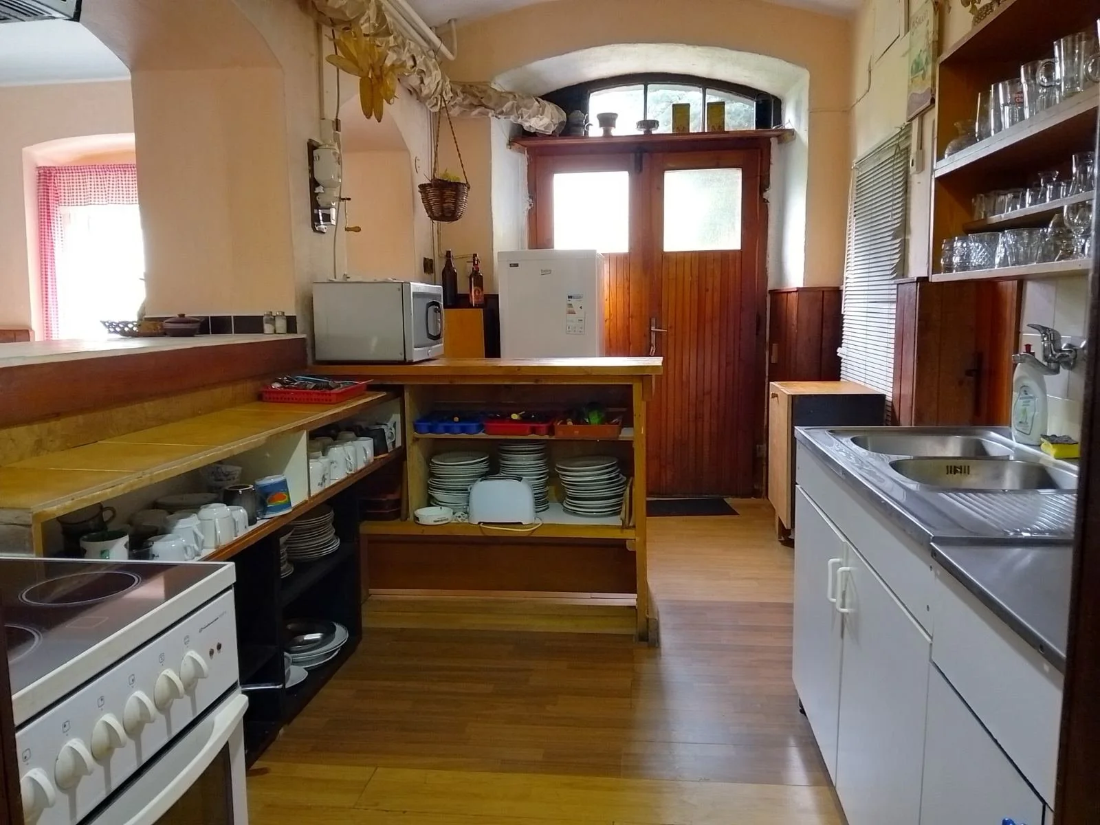 A cozy kitchen with wooden cabinets, a white refrigerator near the door, a microwave on a counter, open shelving with dishes and glasses, a stove in the foreground, and warm natural light coming through a window in the background.