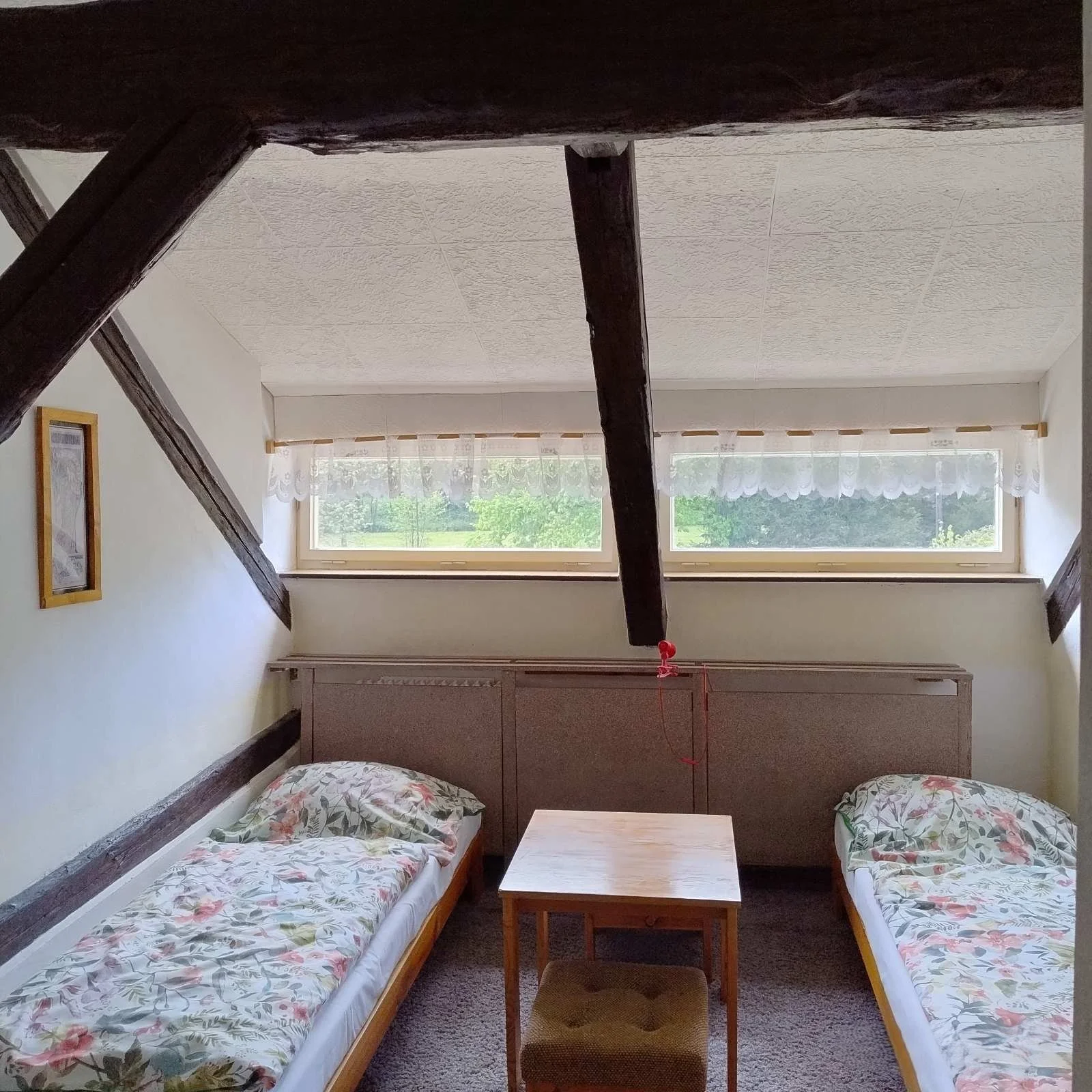 A small bedroom with two twin beds with floral bedding, separated by a wooden table and a small stool. Two large horizontal windows with lace curtains provide natural light, and dark wooden beams cross the ceiling.