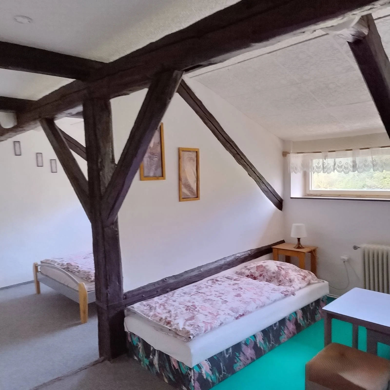 A cozy bedroom with a vintage style, featuring a white bed with floral bedspread, a small wooden side table with a white lamp, a window with lace curtains, and exposed dark wooden beams on the ceiling and wall, with various framed pictures on the wal