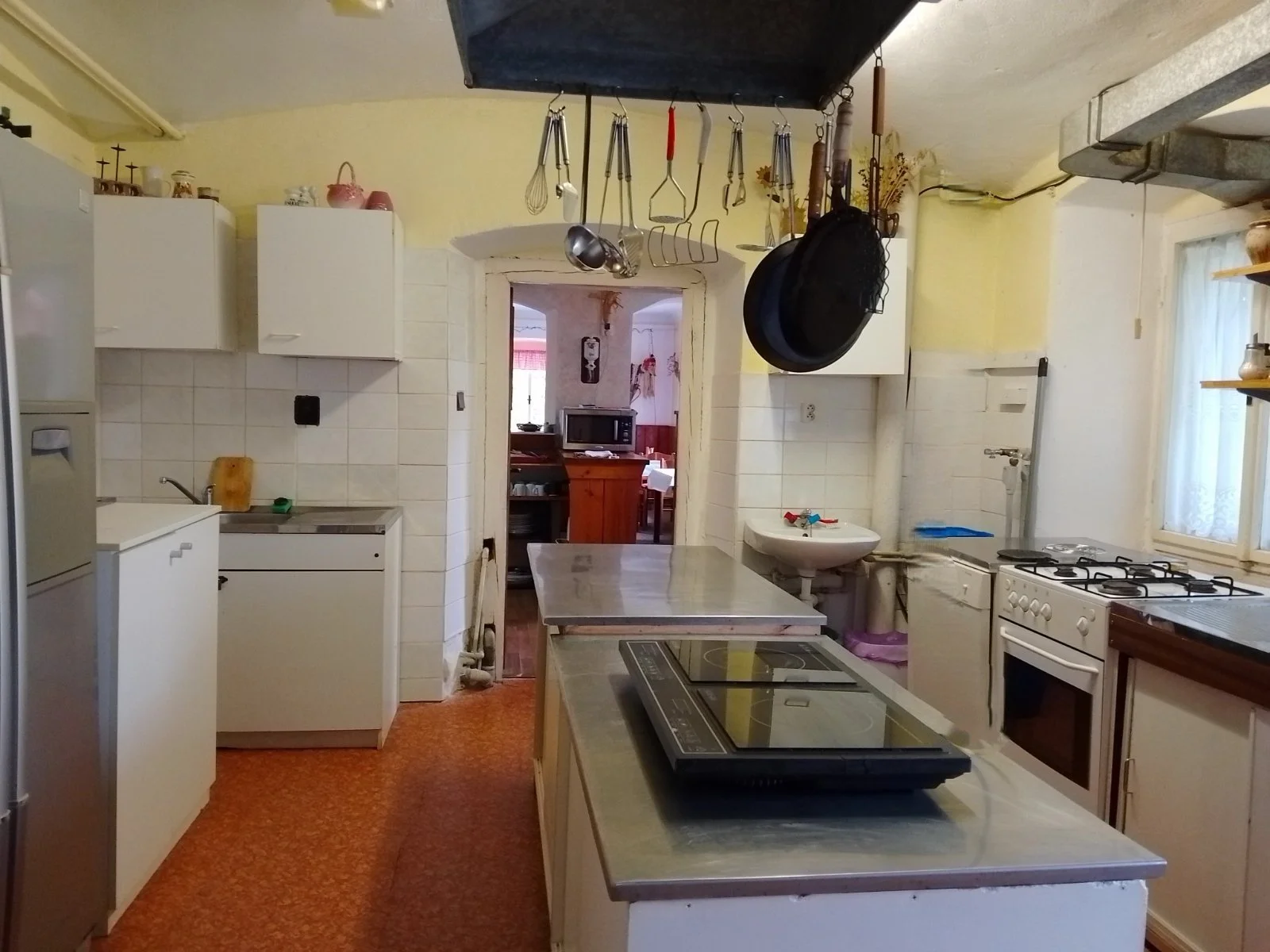 A kitchen with white cabinets, a stove, and various kitchen utensils hanging from the ceiling. There is a microwave in the adjacent room, a sink, and a window with lace curtains. The floor is carpeted in orange.