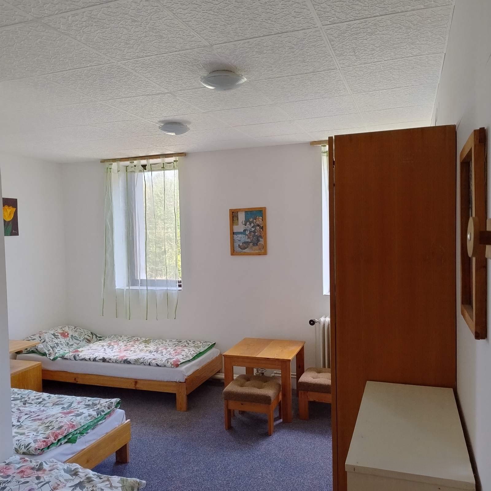 A simple bedroom with three wooden beds with floral bedding, a small wooden table with two chairs, a white dresser, a window with sheer curtains, and framed artwork on the white walls.