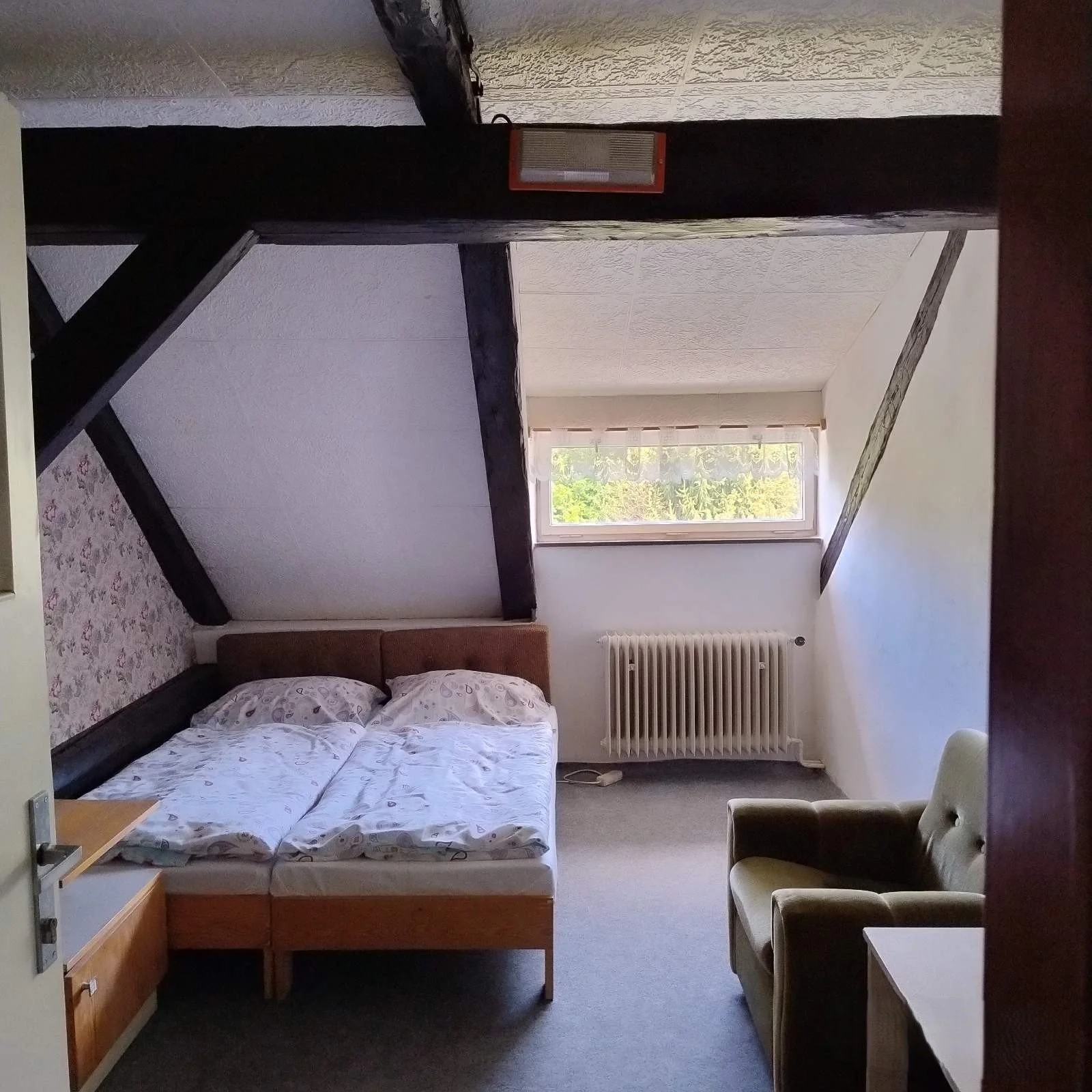 A small bedroom with a bed, a cushioned armchair, and a window with a view of trees outside. The room has white walls, a radiator, and dark wooden beams on the ceiling.