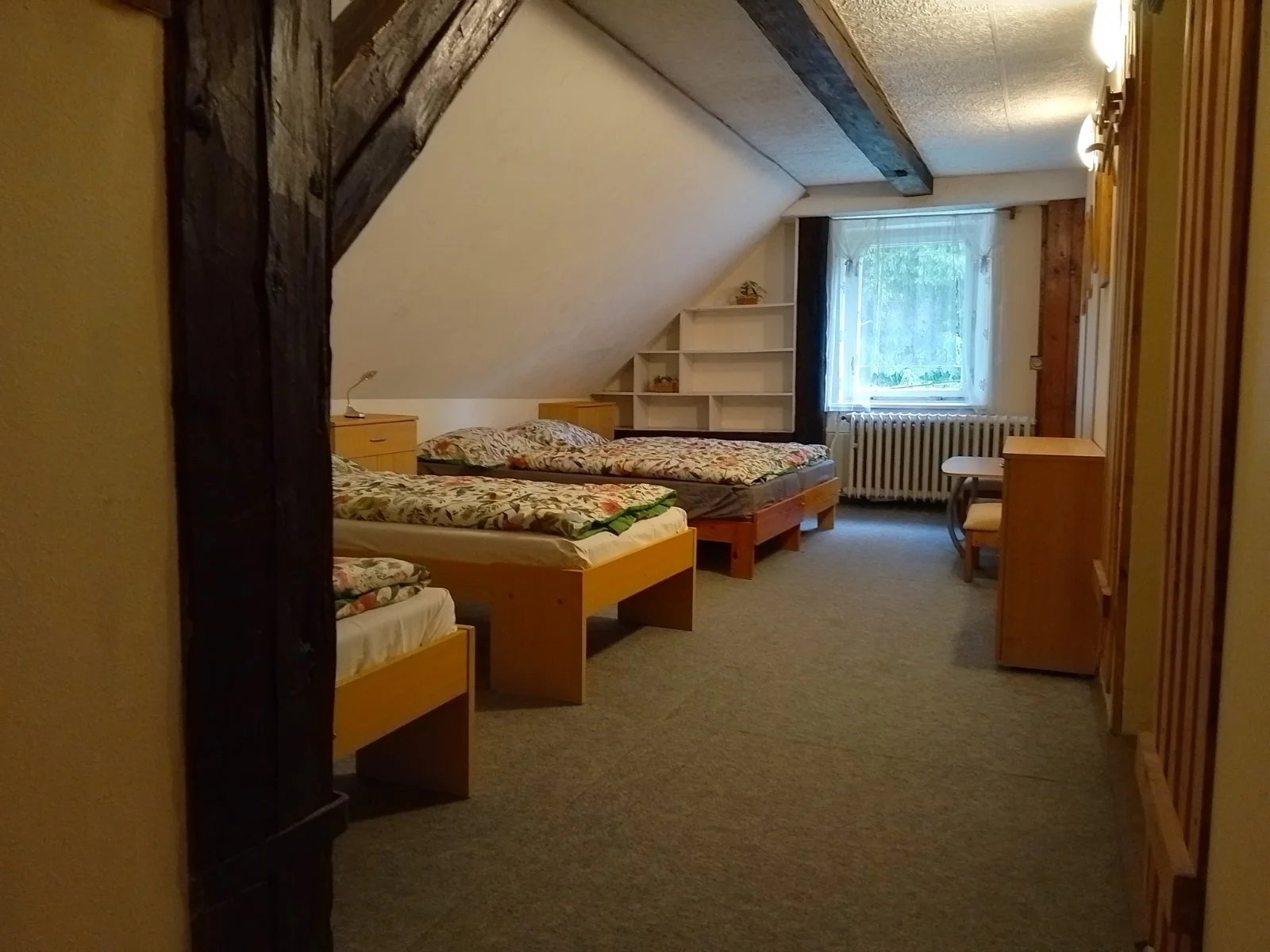 A cozy attic bedroom with three beds, wooden furniture, a window with sheer curtains, and a built-in bookshelf under the sloped ceiling.