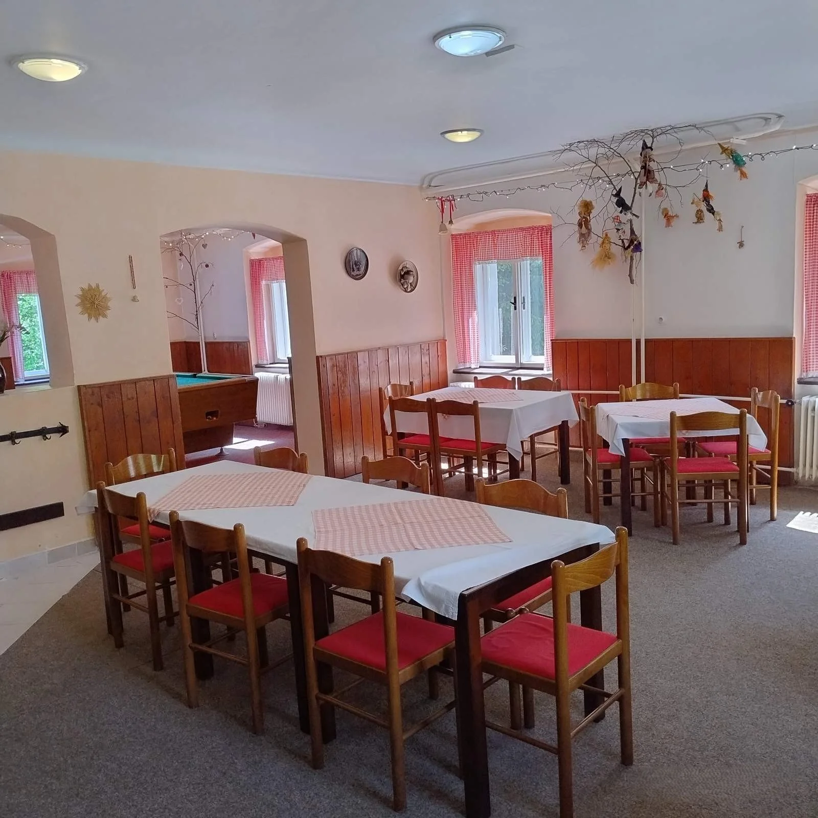 A cozy restaurant with wooden furniture, white tablecloths, and red-cushioned chairs. Decor includes hanging dried flowers, a branch with ornaments, pink curtains, and wall art. Multiple tables are set for dining, with natural light coming through th