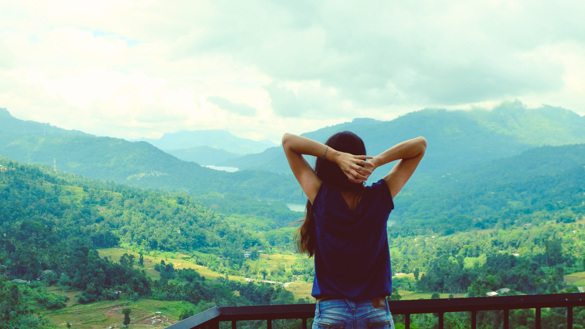 A woman standing on a balcony overlooking a lush green valley with mountains in the background, her arms raised and hands behind her head.