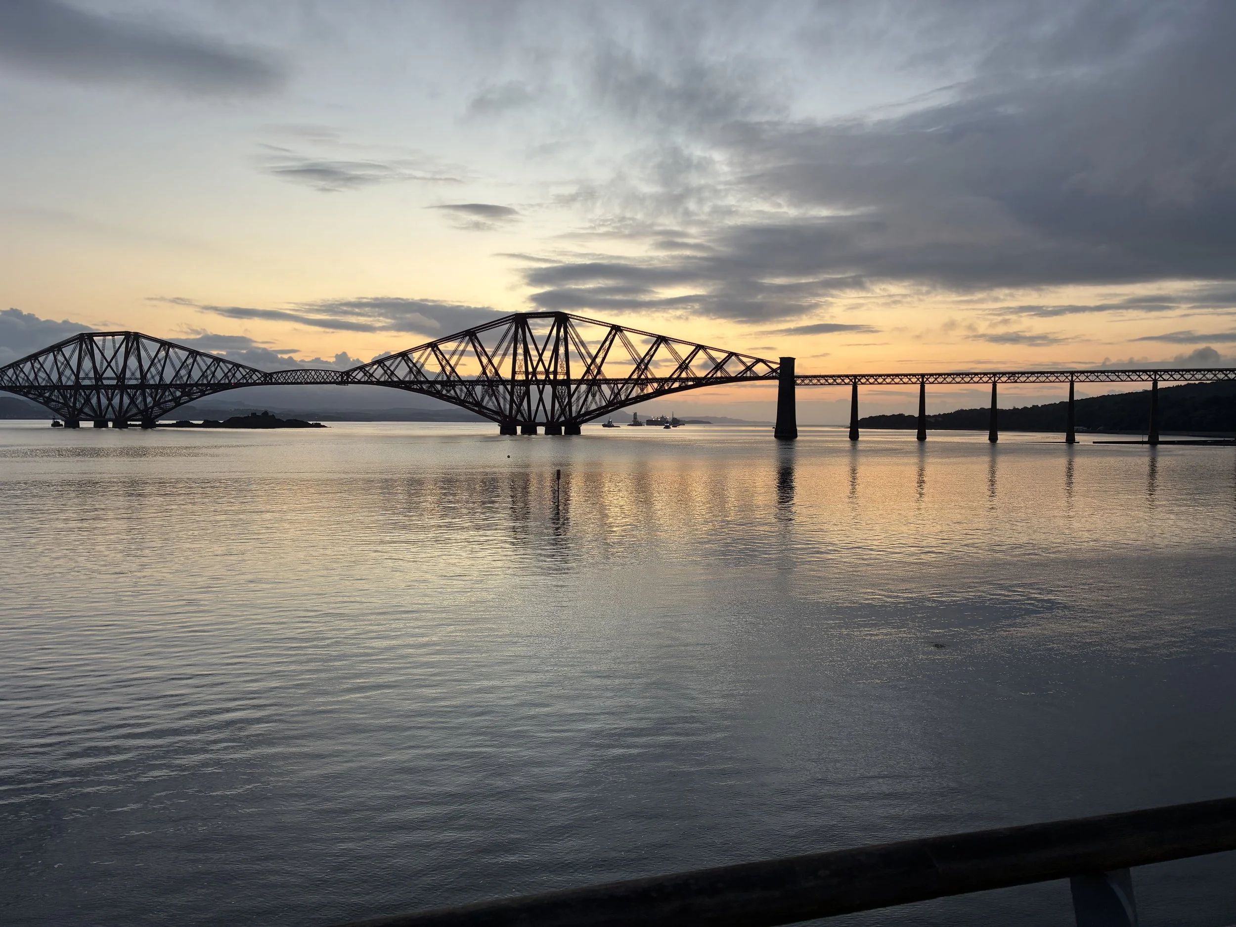A bridge over water at sunset with cloudy sky, calm water reflecting the sky and bridge, and distant ships and land.