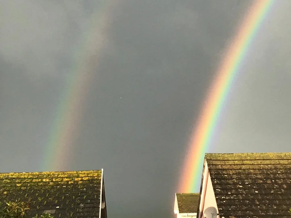 Double rainbow over rooftops with black, shingled roofs under a cloudy sky.
