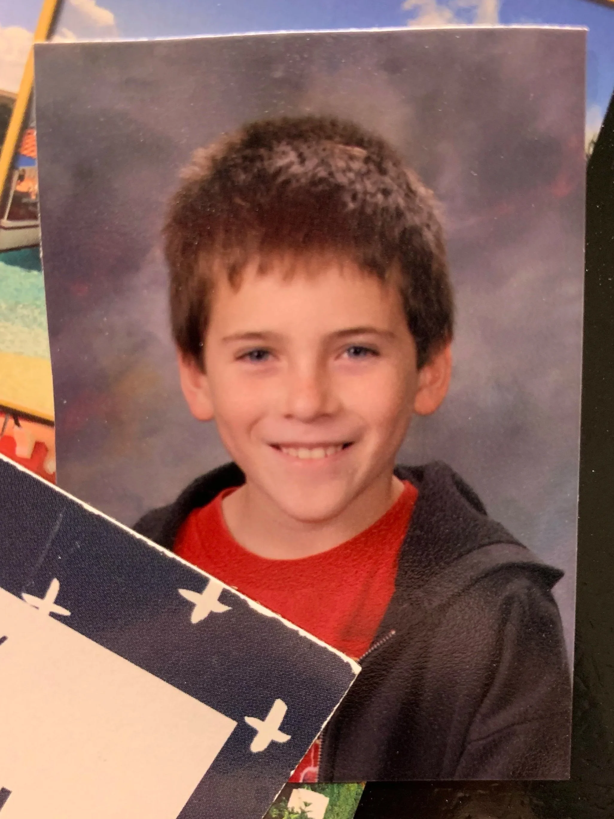 Benjamin Rocha when he was young having short, dark blonde hair while smiling and wearing a red shirt and a black hoodie, with a colorful, soft background.