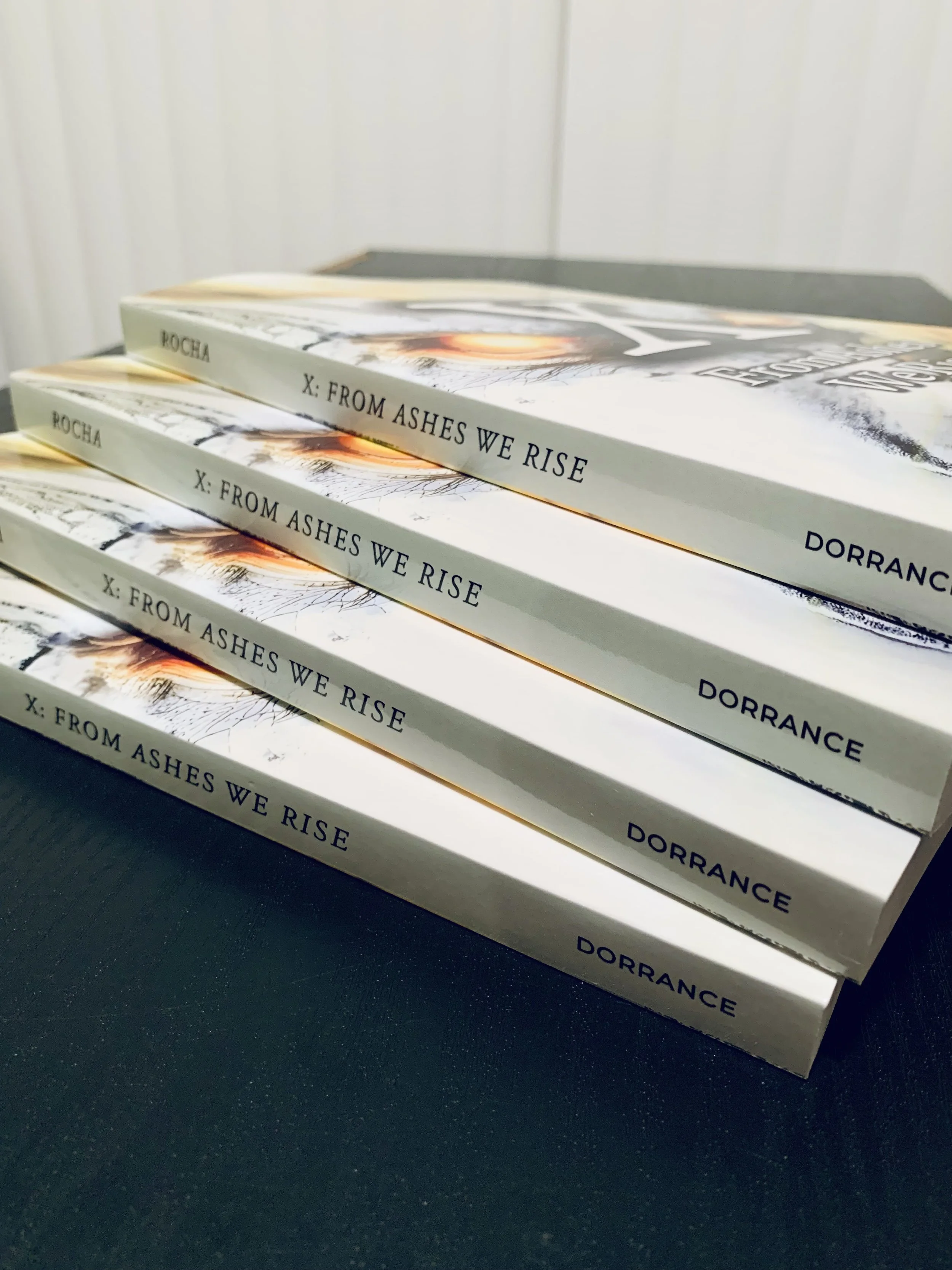 Four copies of the book titled "X: From Ashes We Rise" published by Dorrance Publishing Co and written by Benjamin Rocha stacked on a black surface.