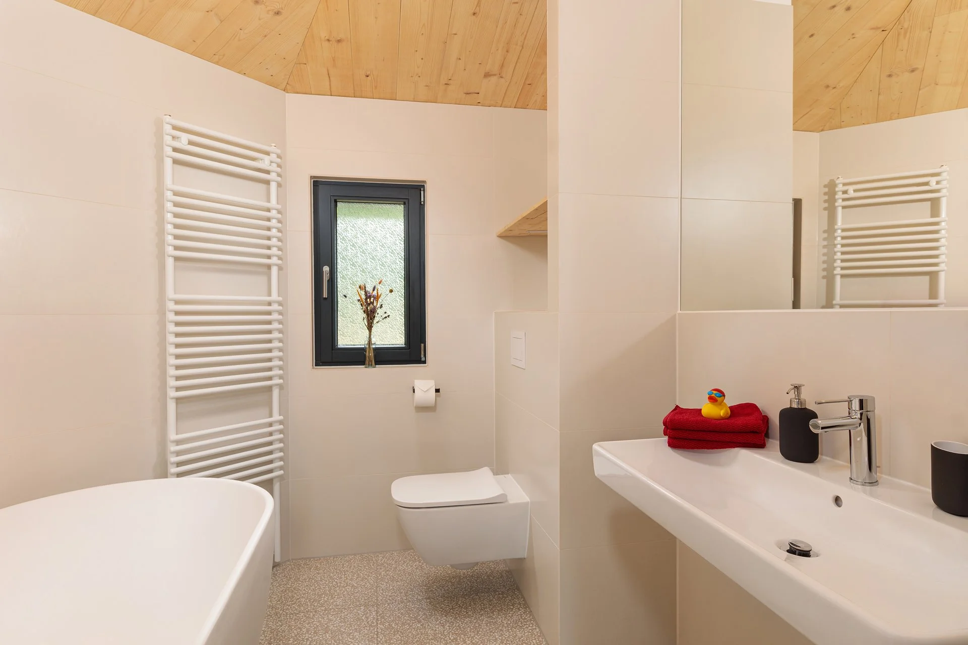 Ein modernes Badezimmer mit weißer Badewanne, Toilette, Waschbecken mit schwarzen und weißen Accessoires, einem Fenster mit Blumen, Holzdecke, weißen Fliesen und Handtüchern.