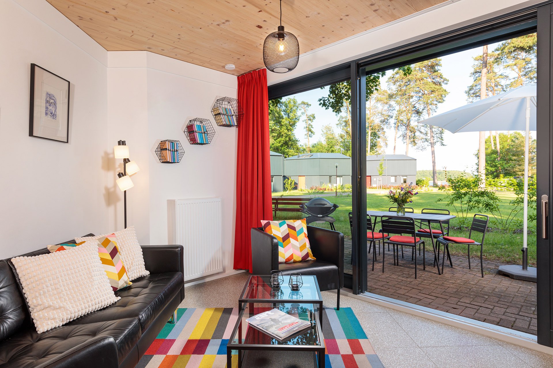 Innenraum eines Wohnzimmers mit schwarzem Ledersofa, bunten Kissen, einem kleinen schwarzen Sessel, einem Glas Couchtisch, buntem Teppich, Wanddekorationen, Stehlampe, roter Vorhang, große Schiebetür öffnet sich auf einen Garten mit Tisch, Stühlen, S