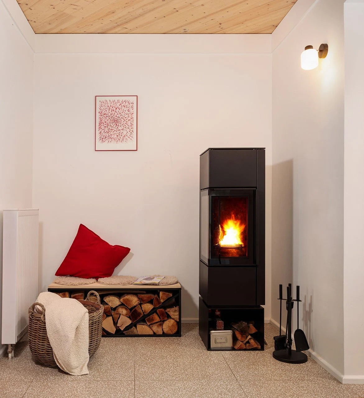 Ein gemütliches Wohnzimmer mit Kamin, Feuer brennt, ein Korb mit Decken, eine Sitzbank mit rotem Kissen und Holzstämmen, Wandbild, Wandlampe, und Kaminbesteck