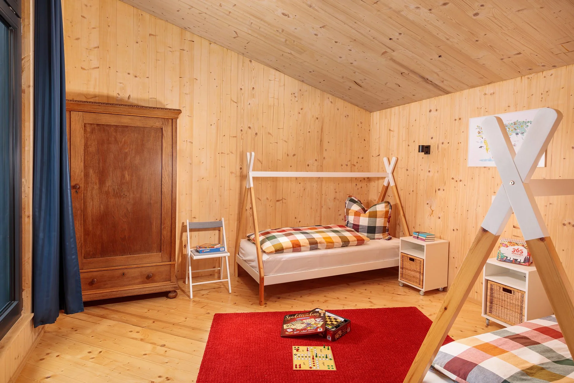Ein Kinderzimmer mit Holzvertäfelung, einem Bett mit Schutzrahmen, einem kleinen weißen Nachttisch, einem roten Teppich, einem Spielbrett, einem Kinderbücherschrank und einem Kleiderschrank aus Holz.