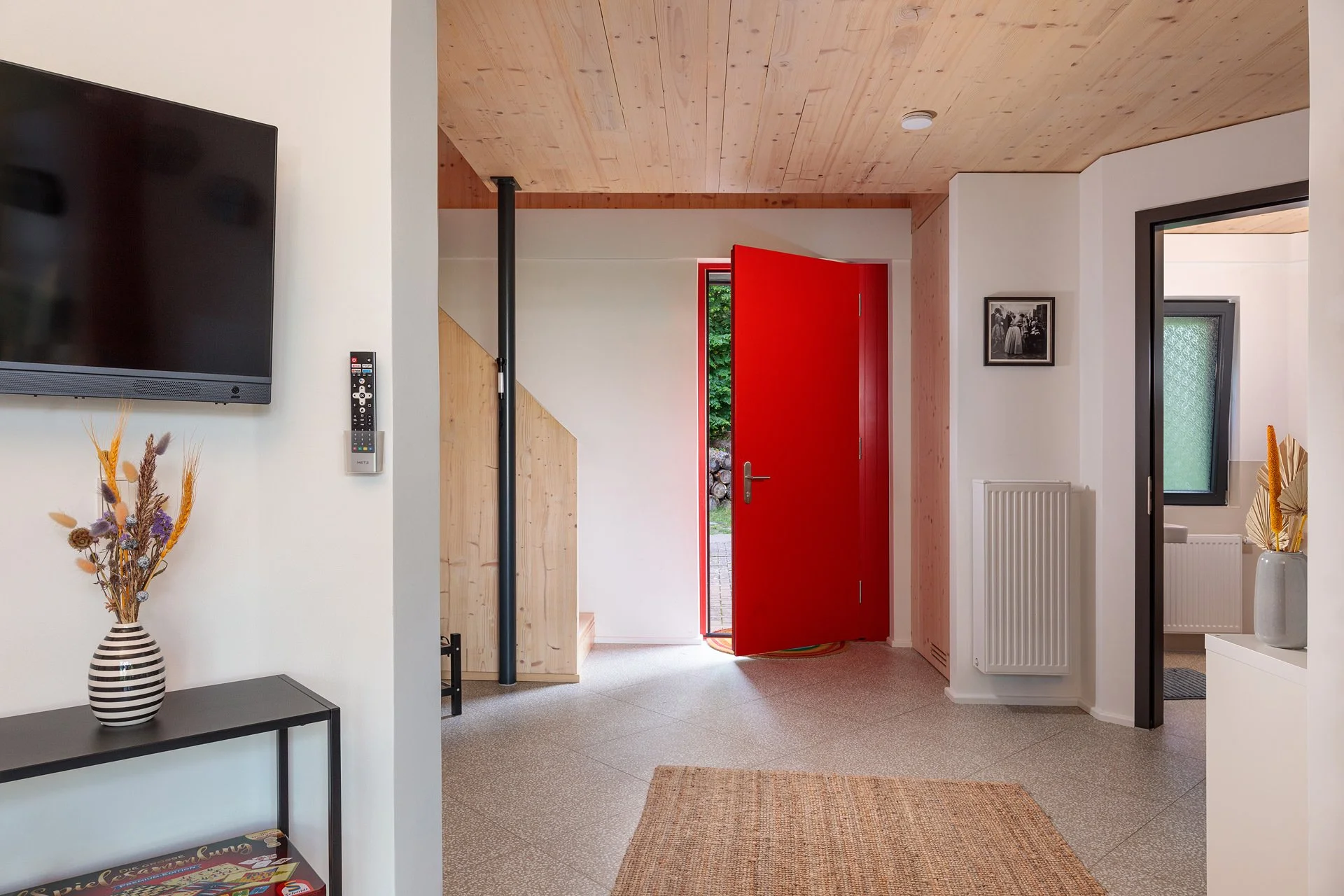 Der Eingangsbereich eines Hauses mit roter Tür, Holzboden und weißen Wänden, mit Dekorationsgegenständen wie einer Vase mit Trockenblumen.