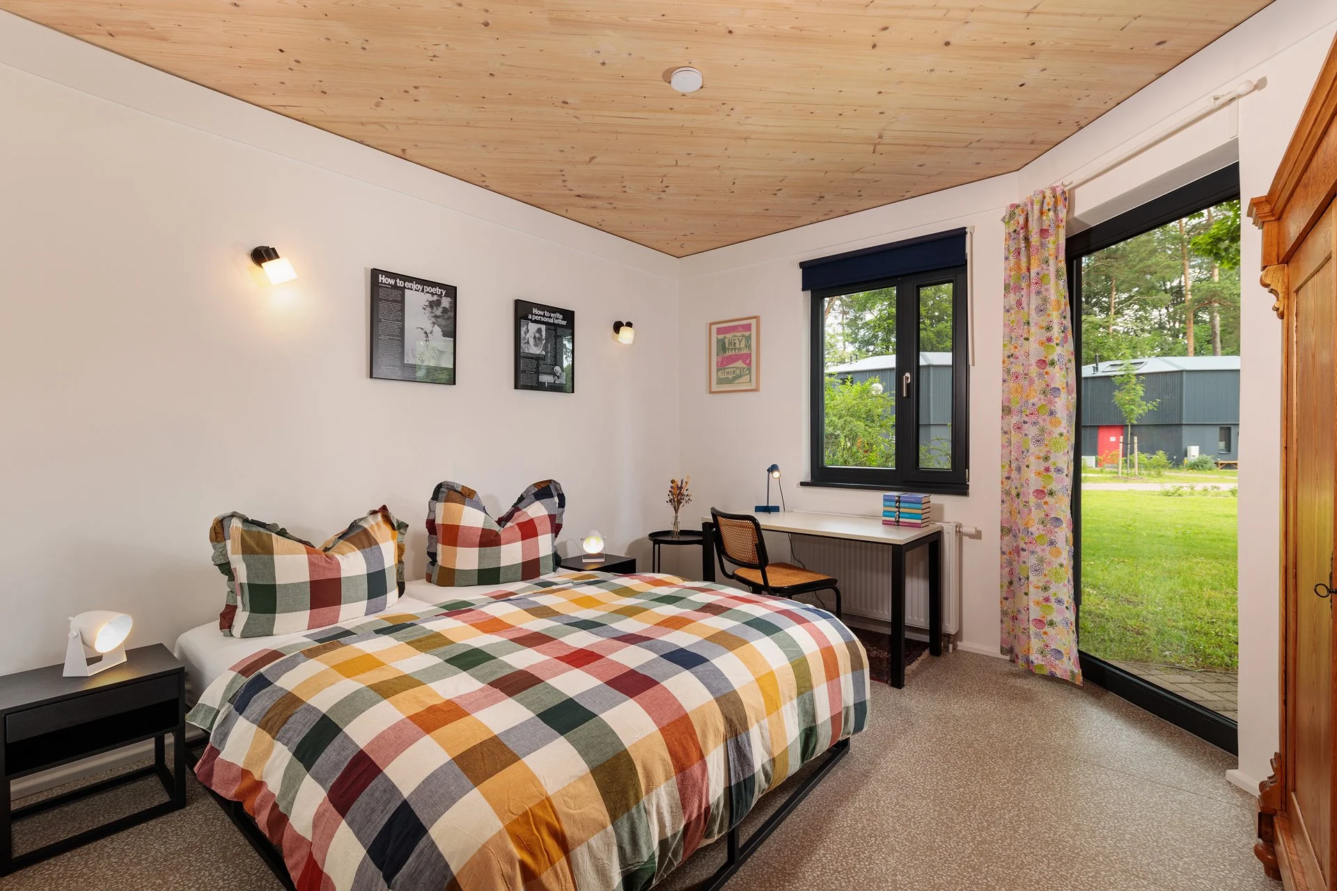 Schlafzimmer mit einem Bett, einer Nachttischlampe, einem Schreibtisch mit Büchern, einem Stuhl, einem Fenster mit Vorhängen und einem offenen Gartenzugang.