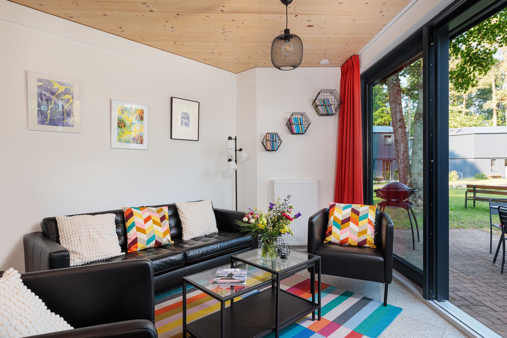 Moderner Wohnzimmer mit schwarzem Sofa, bunten Kissen, Glas-Couchtisch mit Blumen und Zeitschrift, Wandbilder, große Glas-Schiebetür mit Blick auf Garten und Außenbereich