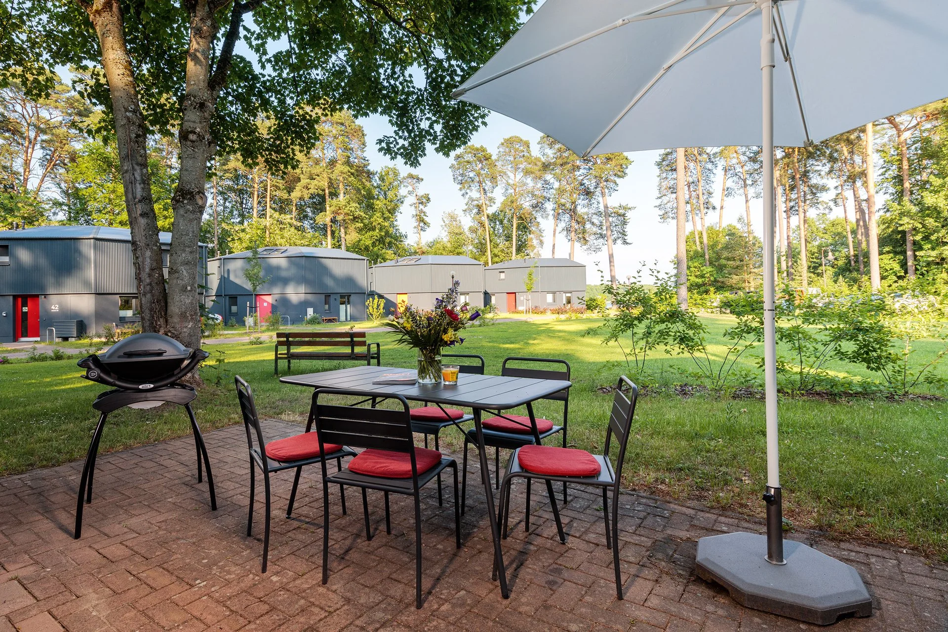 Ein Garten mit einer Sitzgruppe, bestehend aus einem Tisch mit einem Blumenstrauß, sechs Stühlen mit roten Sitzkissen, einem Sonnenschirm, einem Grill, Bäumen und modernen Gebäuden im Hintergrund.