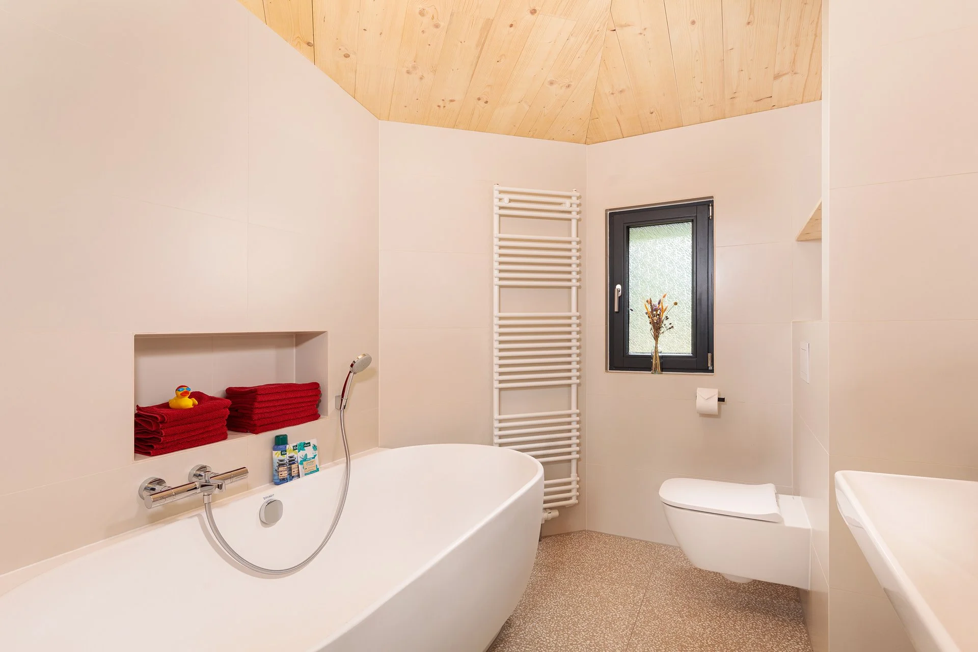 Modernes Badezimmer mit Badewanne, Handtüchern, Fenster und Toilette in neutralen Farben, mit Holzdecke