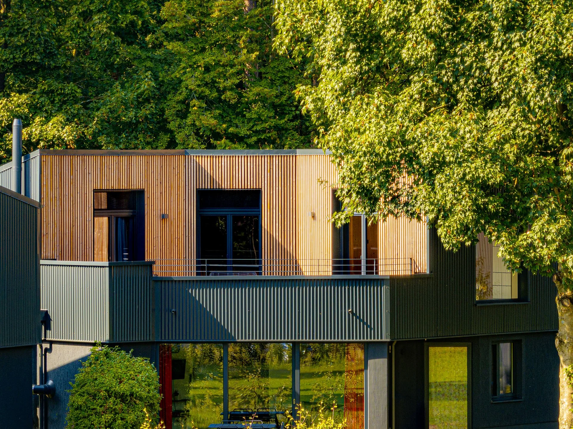 Modernes zweistöckiges Haus mit Veranda, große Fenster, Holzvertäfelung und umgeben von Bäumen.