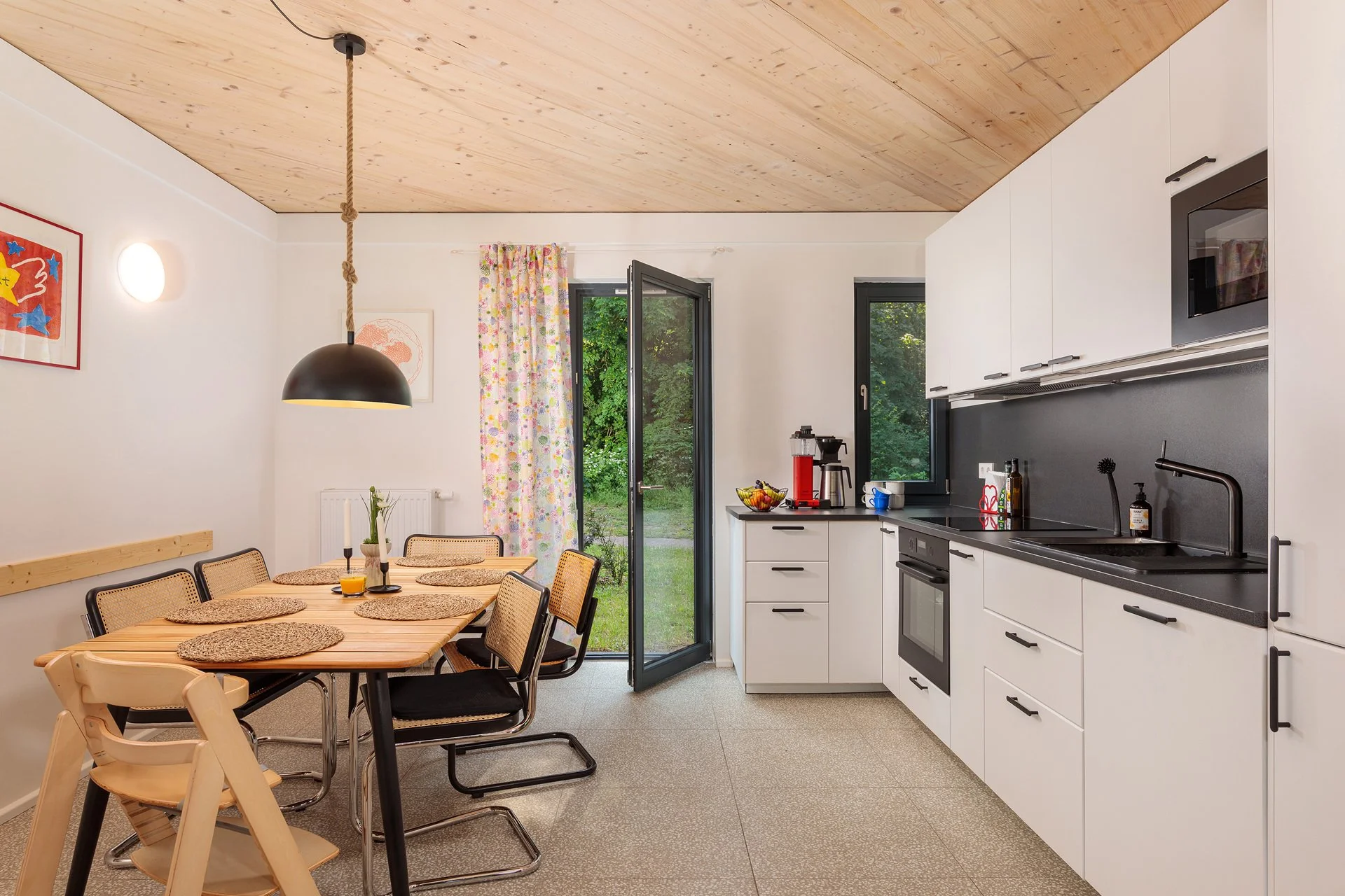 Küche mit Essbereich, weißem Küchenblock, schwarzen Arbeitsflächen, Ofen, Kaffeemaschine, Esszimmer mit Holztisch und schwarzen Stühlen, Fenster und Tür zur grünen Außenfläche, Holzdecke, bunte Vorhänge, Wanddeko und Beleuchtung.