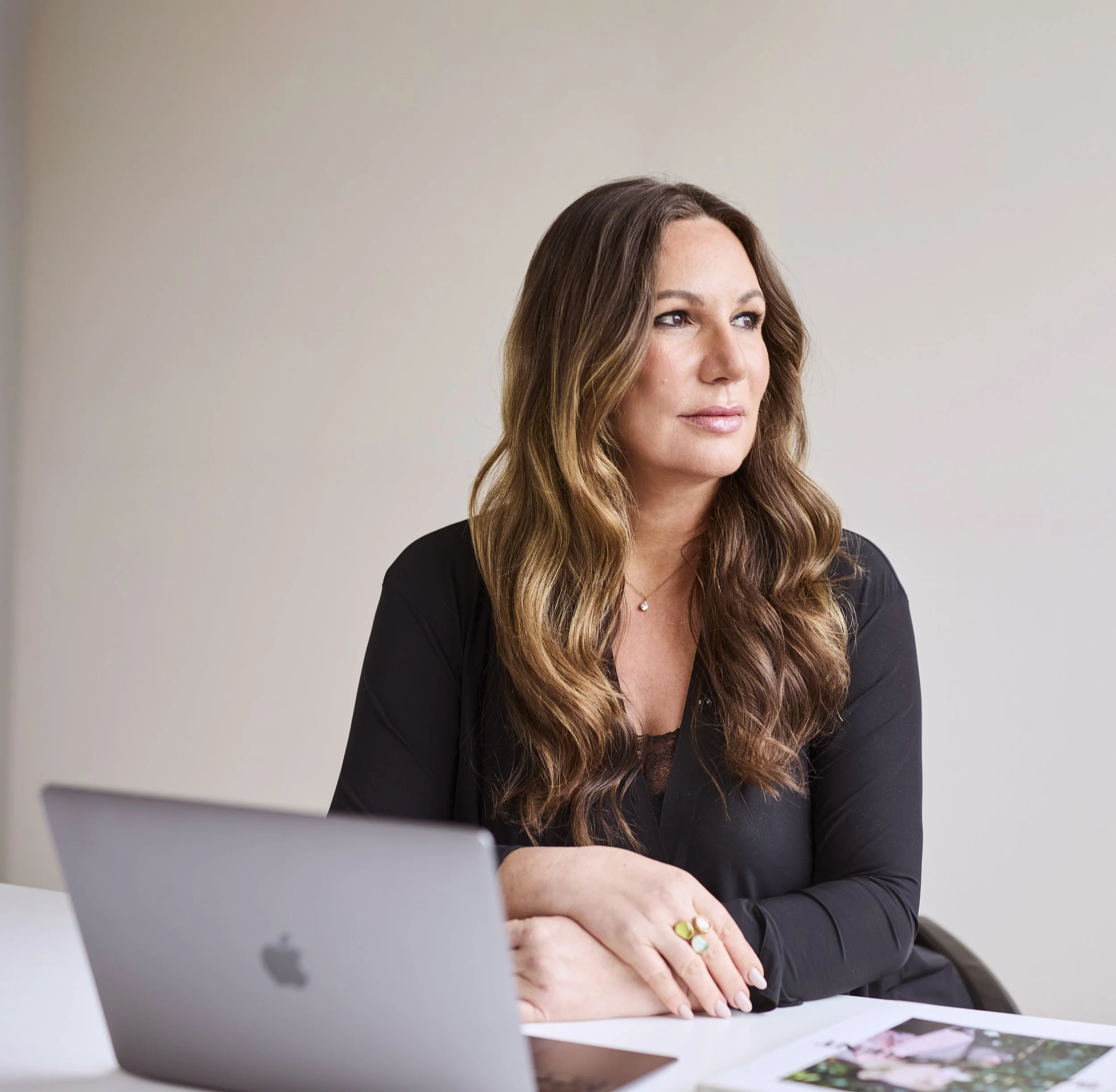 Frau mit langen, lockigen braunen Haar, sitzt an einem Tisch mit einem MacBook, blickt nach rechts, trägt schwarze Kleidung und eine kleine Halskette.