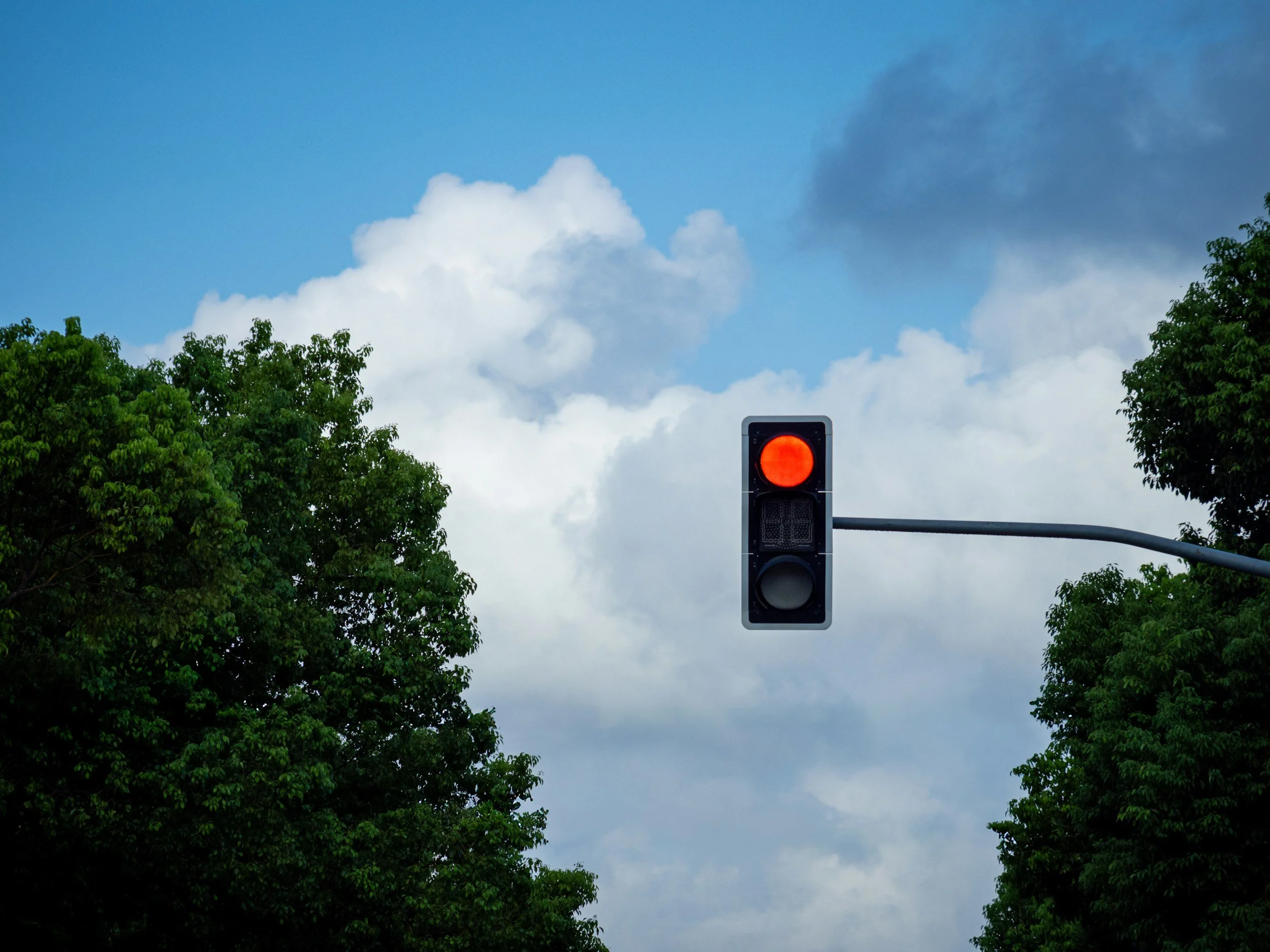 Ampel mit rotem Licht, umgeben von grünen Bäumen und einem Himmel mit Wolken.