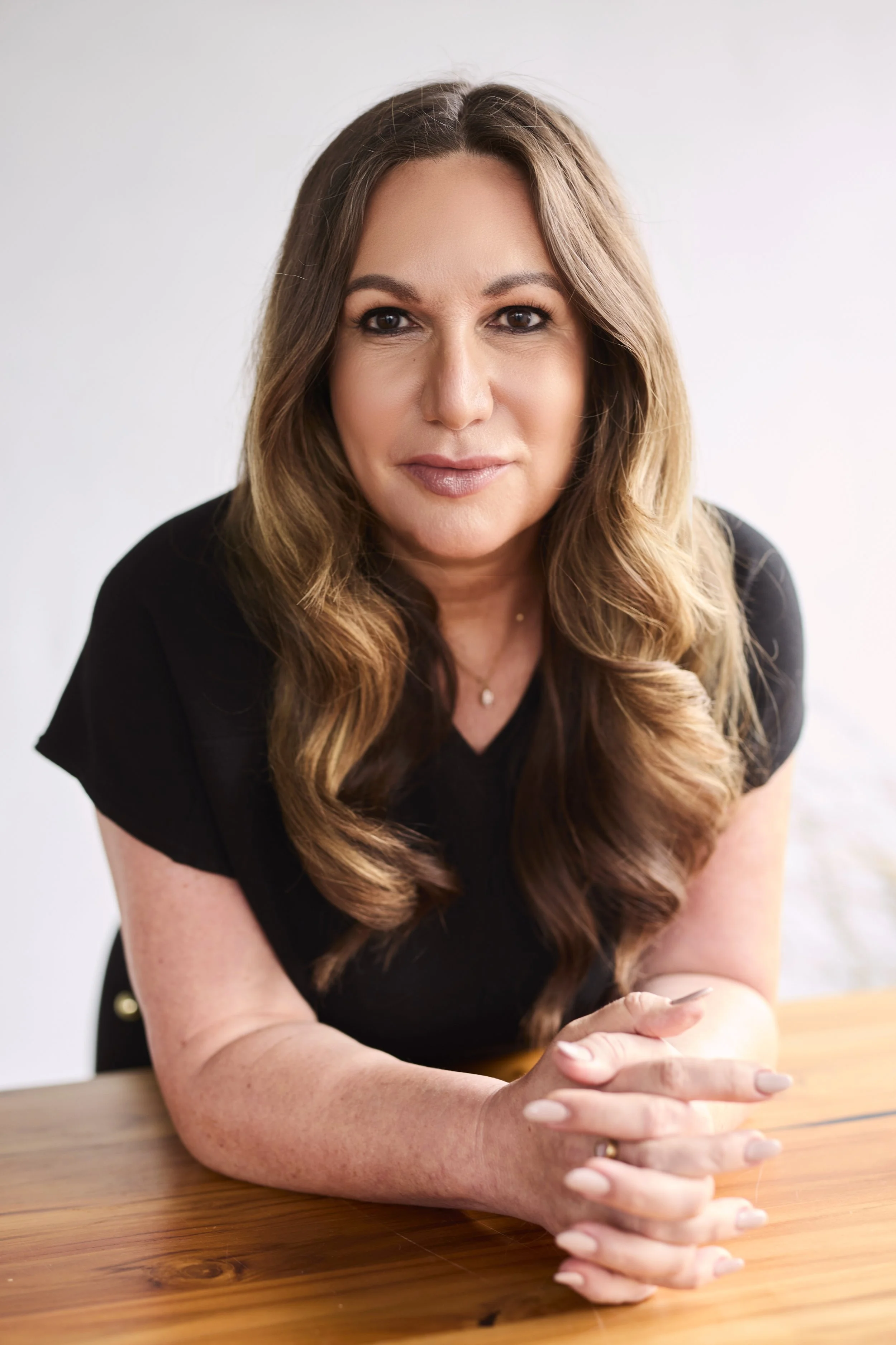 Eine Frau sitzt an einem Holztisch, trägt schwarzes Oberteil, hat lange, lockige braune Haare und schaut in die Kamera.