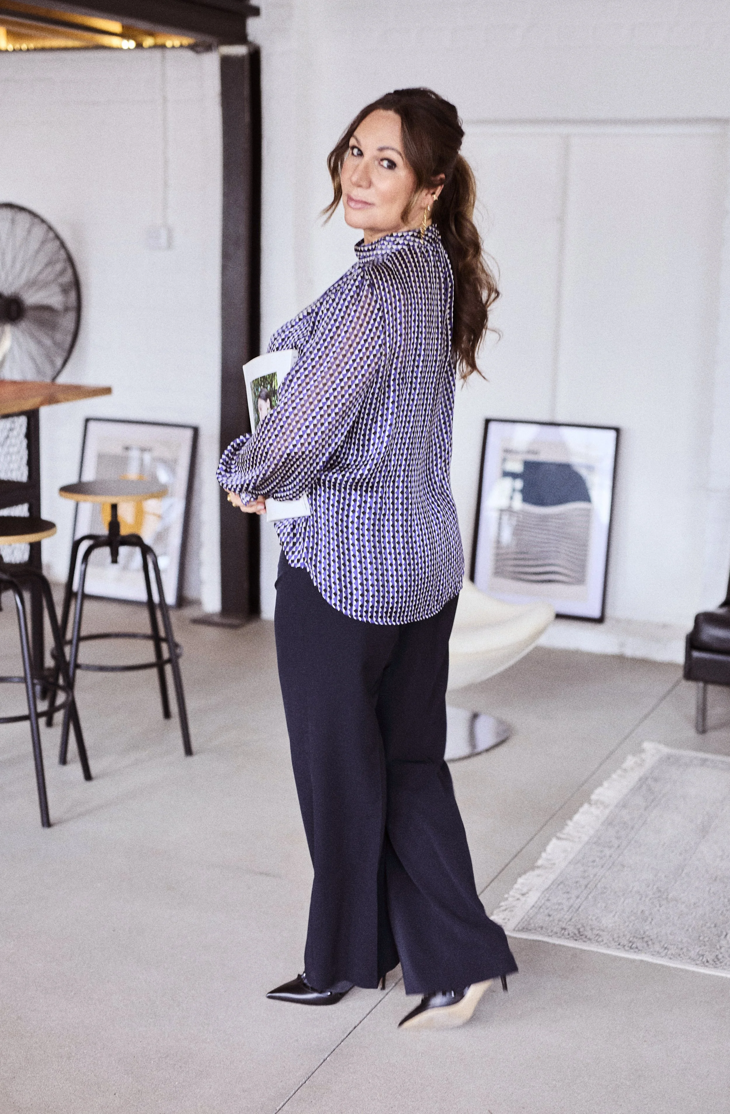 Frau mit braunen, gewellten Haaren trägt eine blusenkreuzartige Bluse mit Leopardenmuster und schwarze Hosen, hält eine Zeitschrift in den Armen, steht in einem modernen Wohnzimmer mit Kunstwerken, Sessel und Stehtisch.