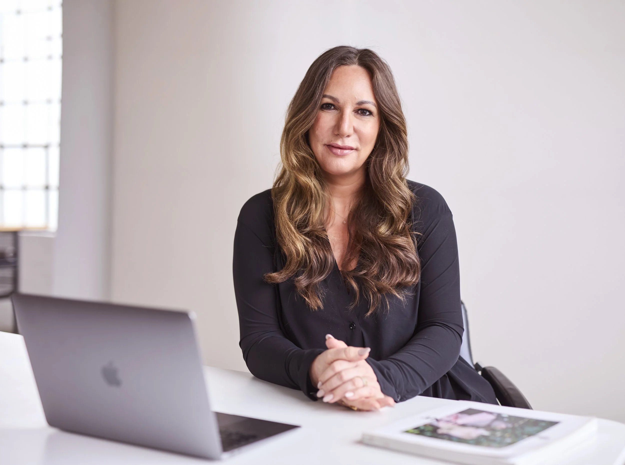 Eine Frau sitzt an einem weißen Schreibtisch vor einem Laptop, umgeben von Akten und Zeitschriften in einem modernen Büro.