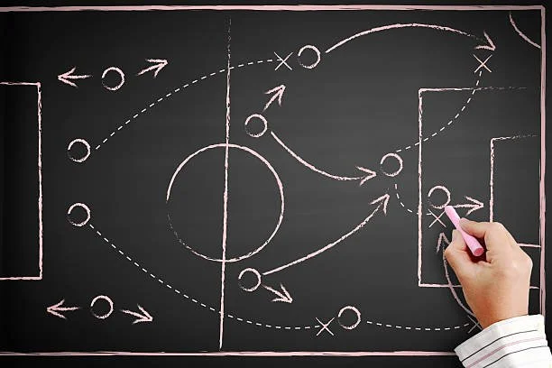 A person drawing soccer strategy plays on a black chalkboard with pink chalk.