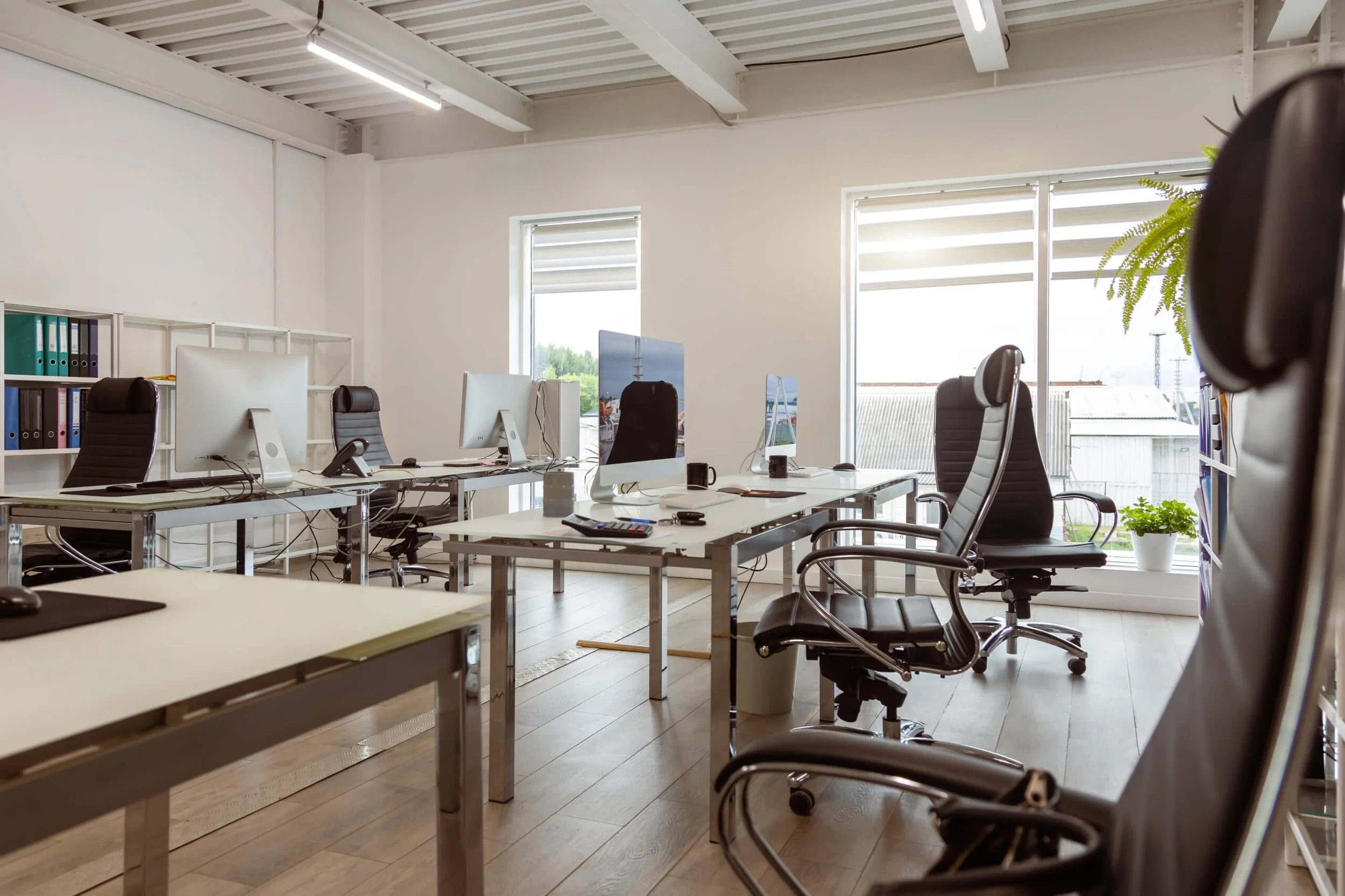 Empty office with white walls, large windows, black ergonomic chairs, desks with computers, and office supplies, sunlight coming through the windows.