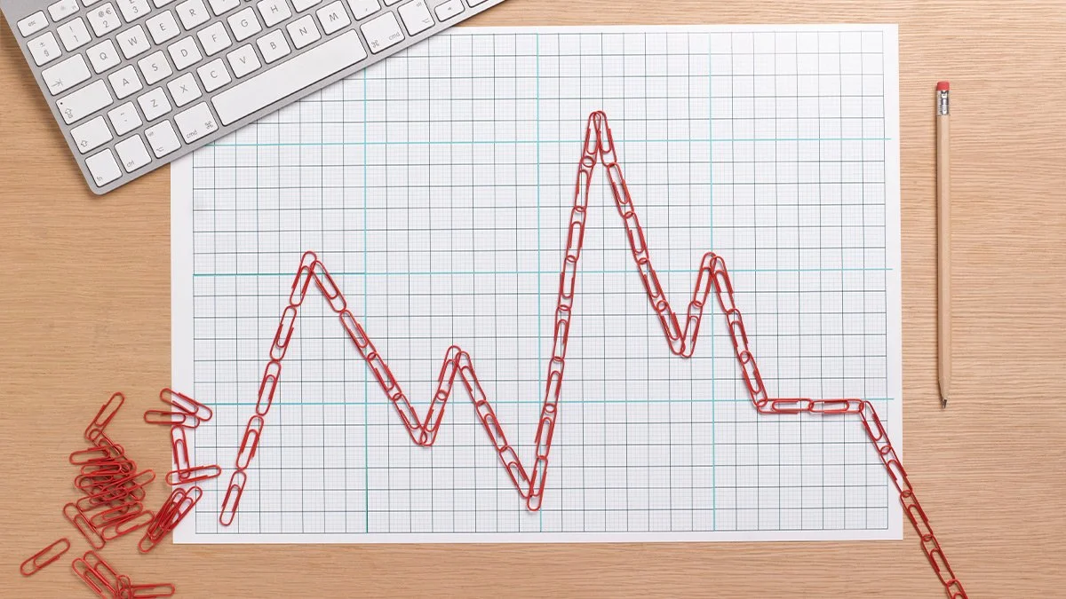 Paper with a printed grid, red paper clips arranged to form a zigzag line resembling a business update's line chart, surrounding a computer keyboard and a pencil on a wooden desk.
