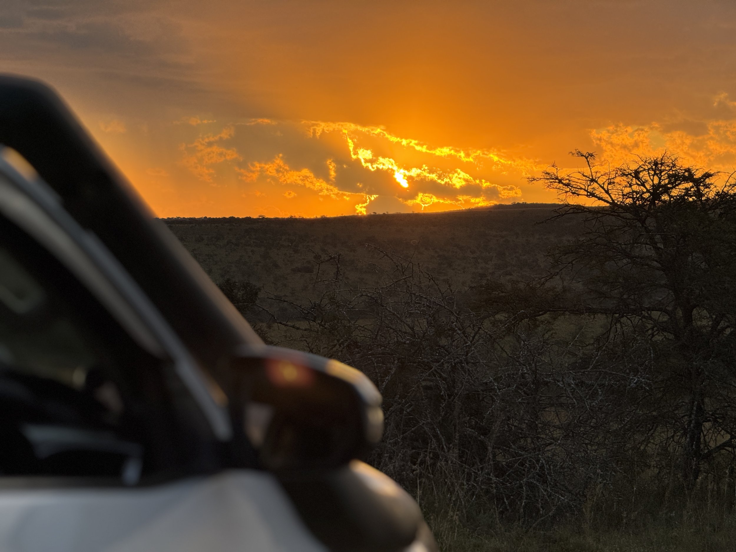 A sunset over dry hills with silhouetted trees and clouds, captured from inside a vehicle