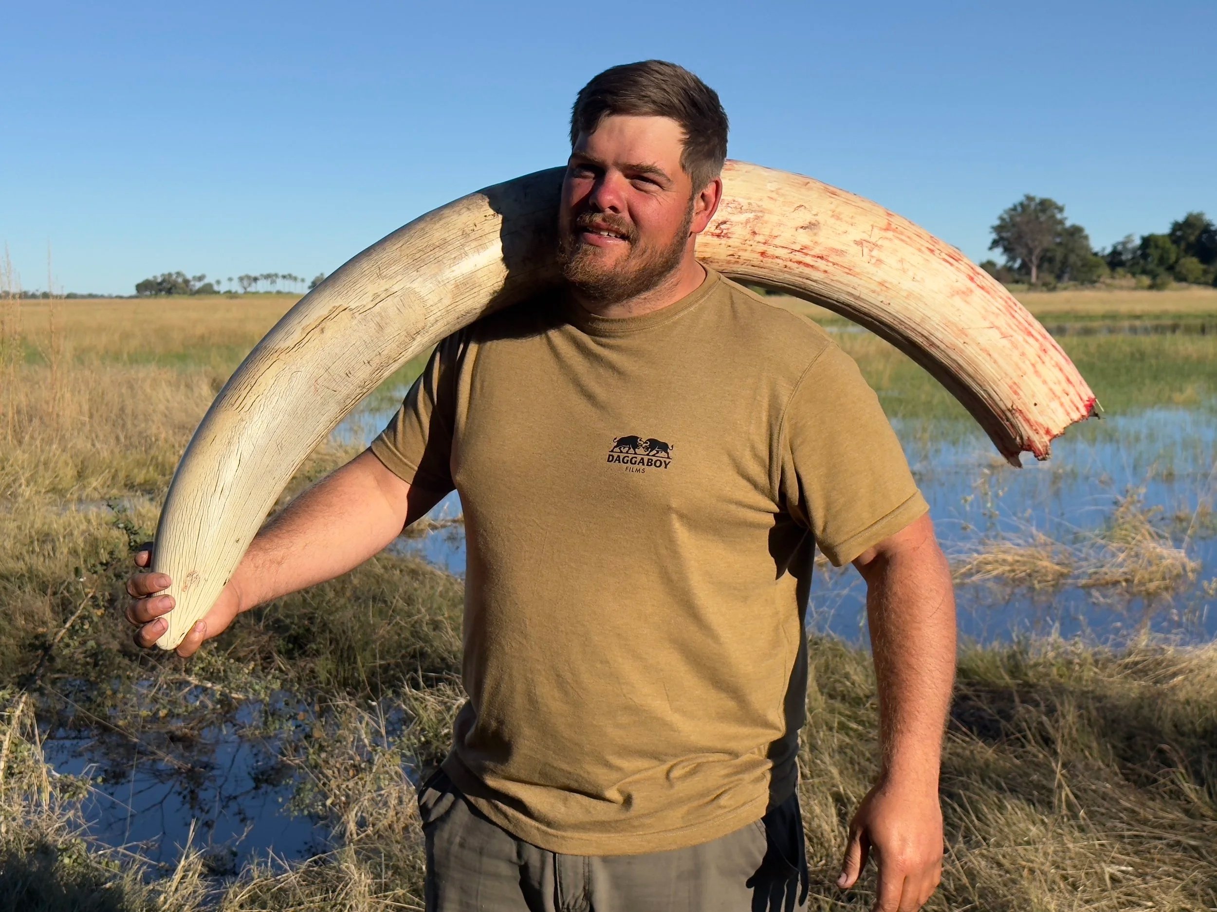 A man with a beard and short hair stands outdoors, holding a large tusk over his shoulder. The background features marshland with water, grass, and scattered trees, under a clear blue sky.
