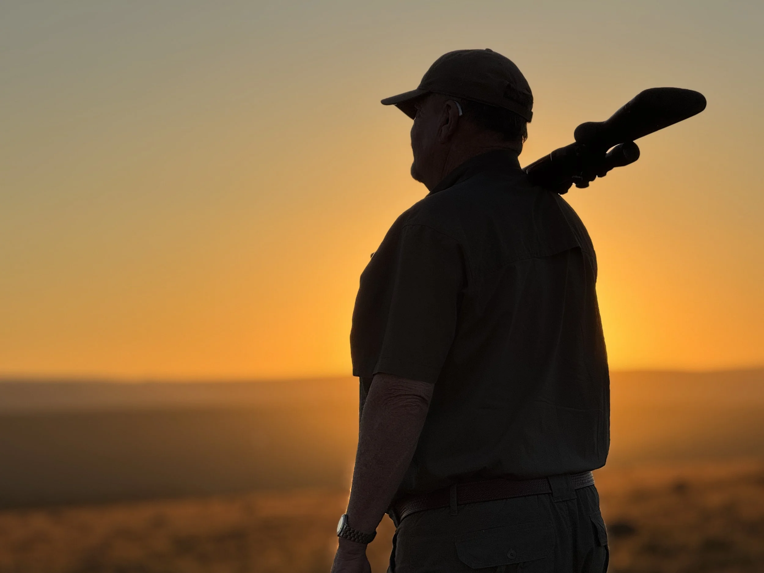 Silhouette of a man carrying a rifle on his shoulder, standing outdoors during sunset.