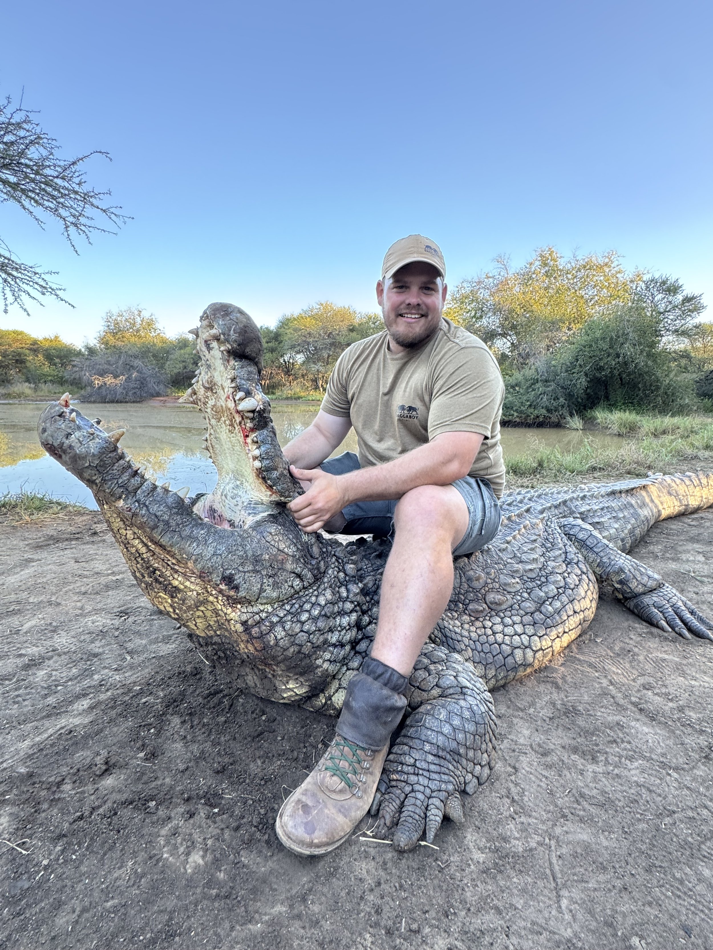 A man sitting on a large crocodile by a riverbank, holding open its mouth, smiling at the camera, with trees and water in the background.