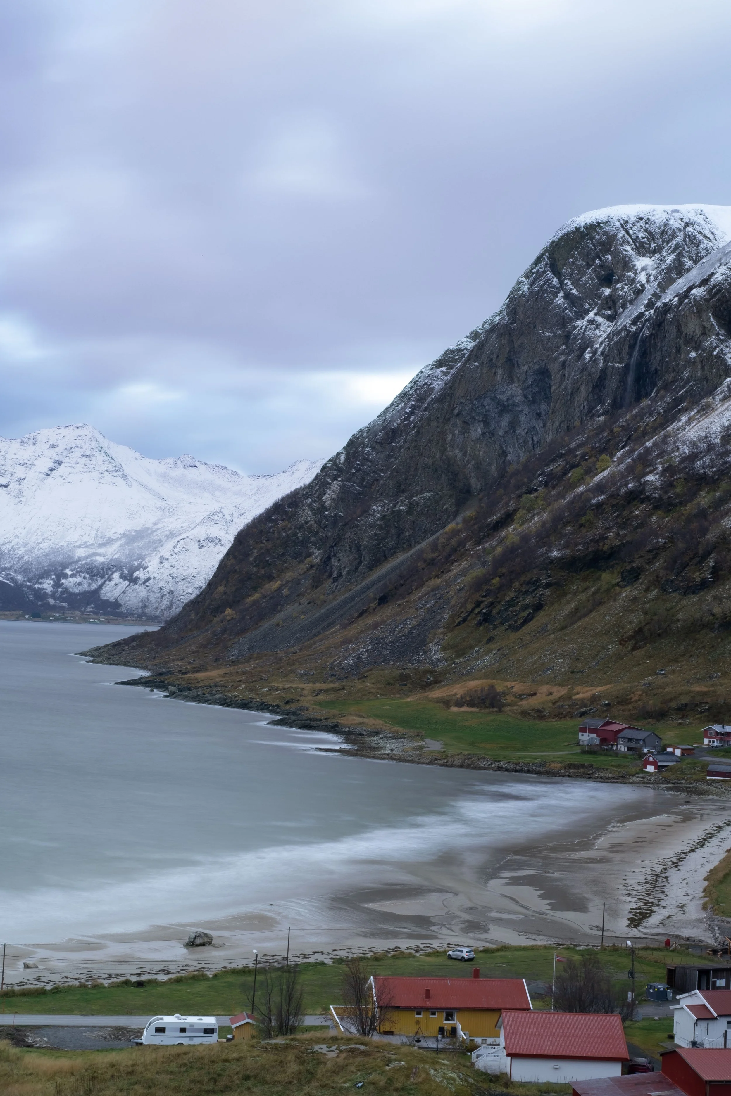A coastal landscape showing snow-capped mountains, a shoreline, a few houses, and a cloudy sky.