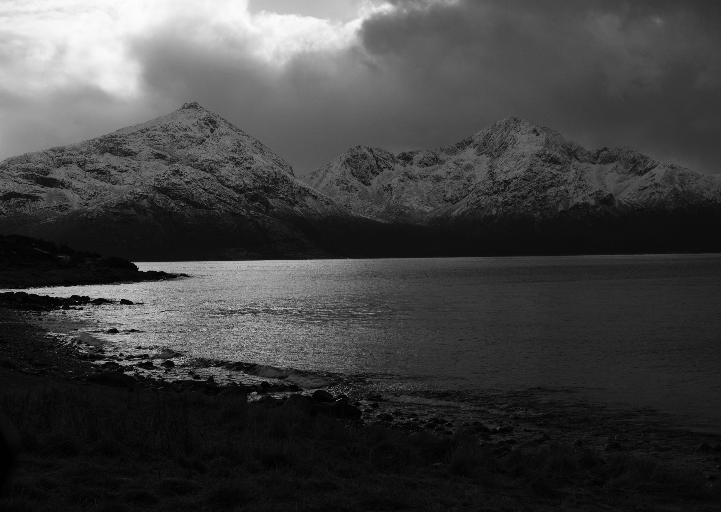 A black and white photo of a lake with mountains in the background and cloudy sky.