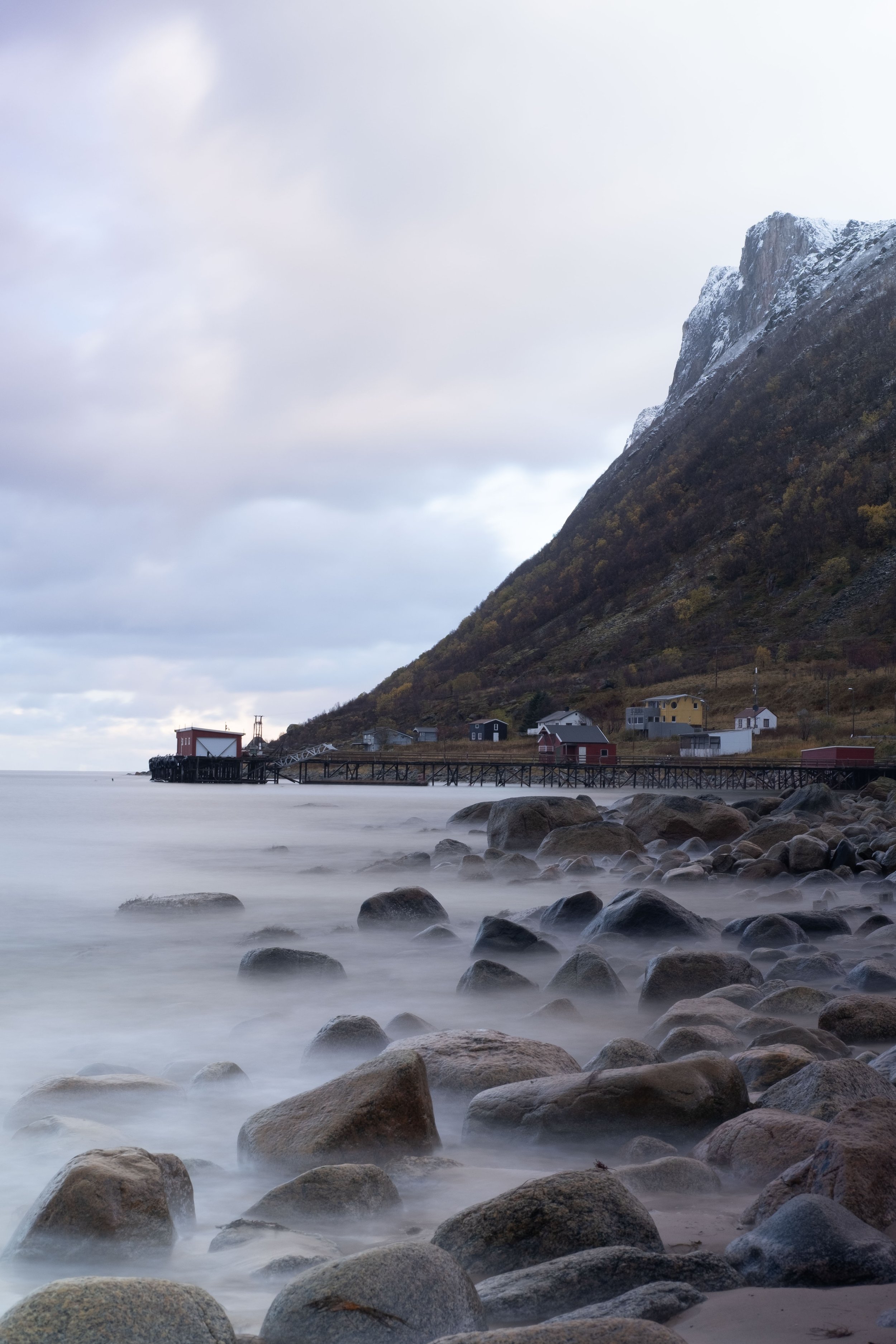 Scenic coastal view with rocky shoreline, calm water, a pier, and colorful houses at the base of a snow-capped mountain under cloudy sky.