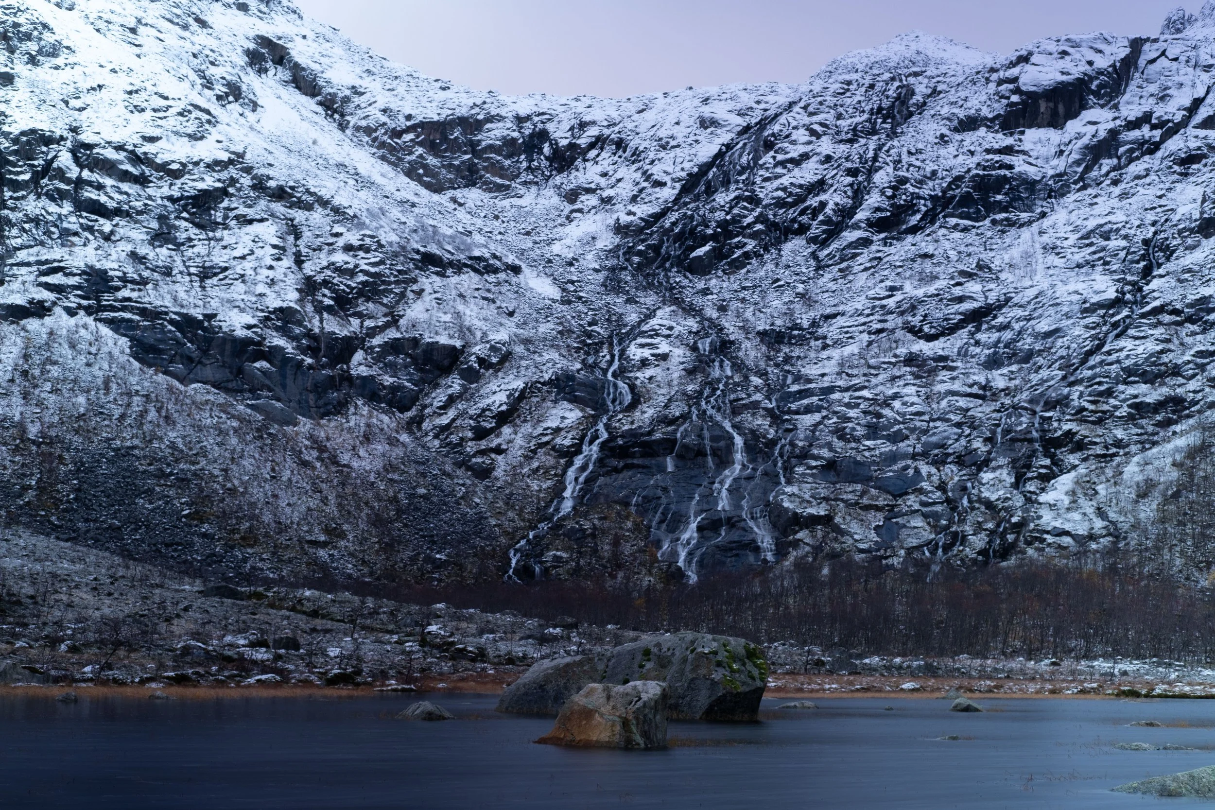 Snow-covered mountain with a waterfall cascading down, a partially frozen lake with rocks in the foreground.
