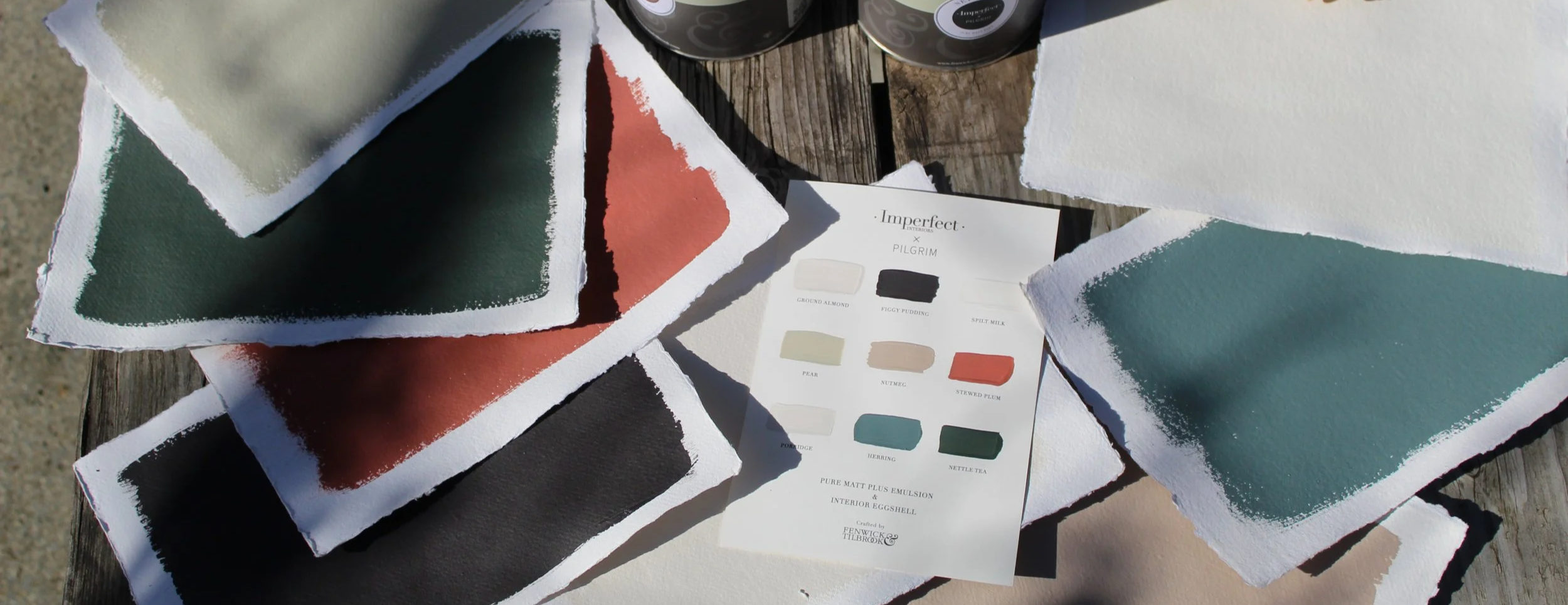 Imperfect Interiors x Pilgrim Paint range swatches in various shades of green, coral,  and beige on textured paper, with colour palette chart and cans of paint on weathered wooden surface.