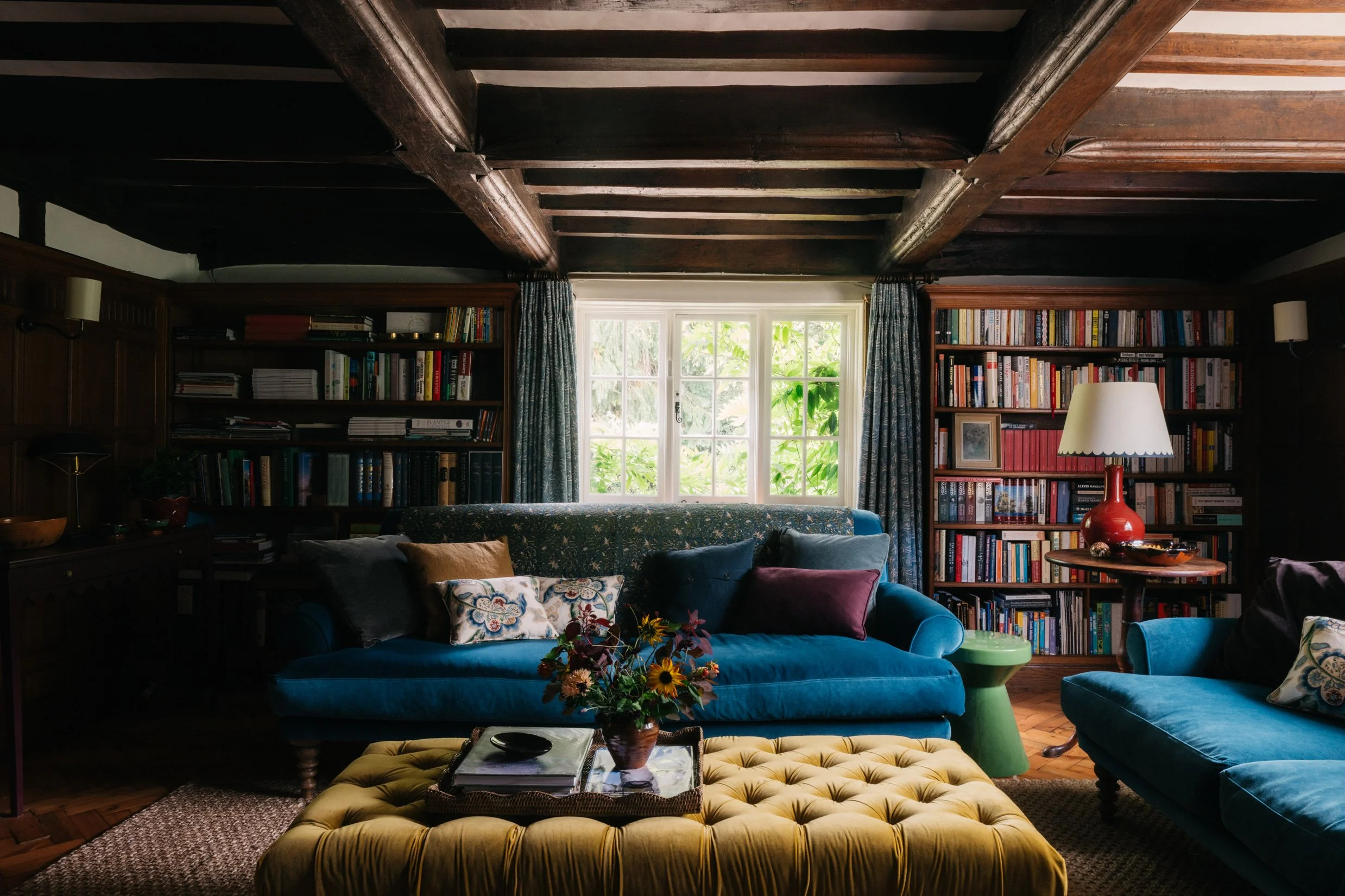 Living room designed by Imperfect Interiors with wooden ceiling beams, blue velvet sofas, a yellow buttoned ottoman with a tray and flowers, bookshelves filled with books, a window with blue curtains, and a table with a red lamp.
