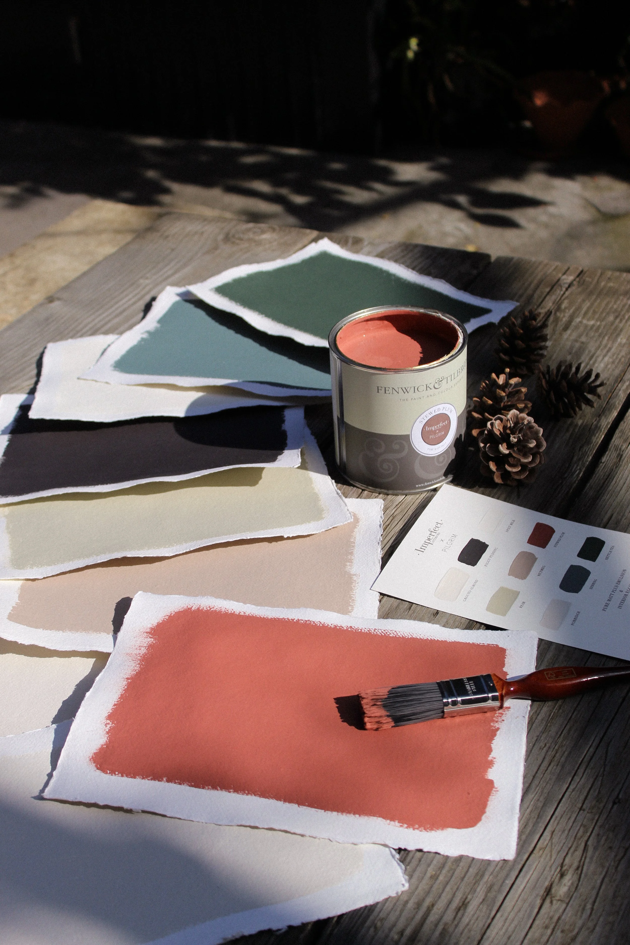 Imperfect Interiors x Pilgrim Paint swatches, a can of paint, a paintbrush, pinecones, and a colour sample sheet arranged on a wooden table outdoors.