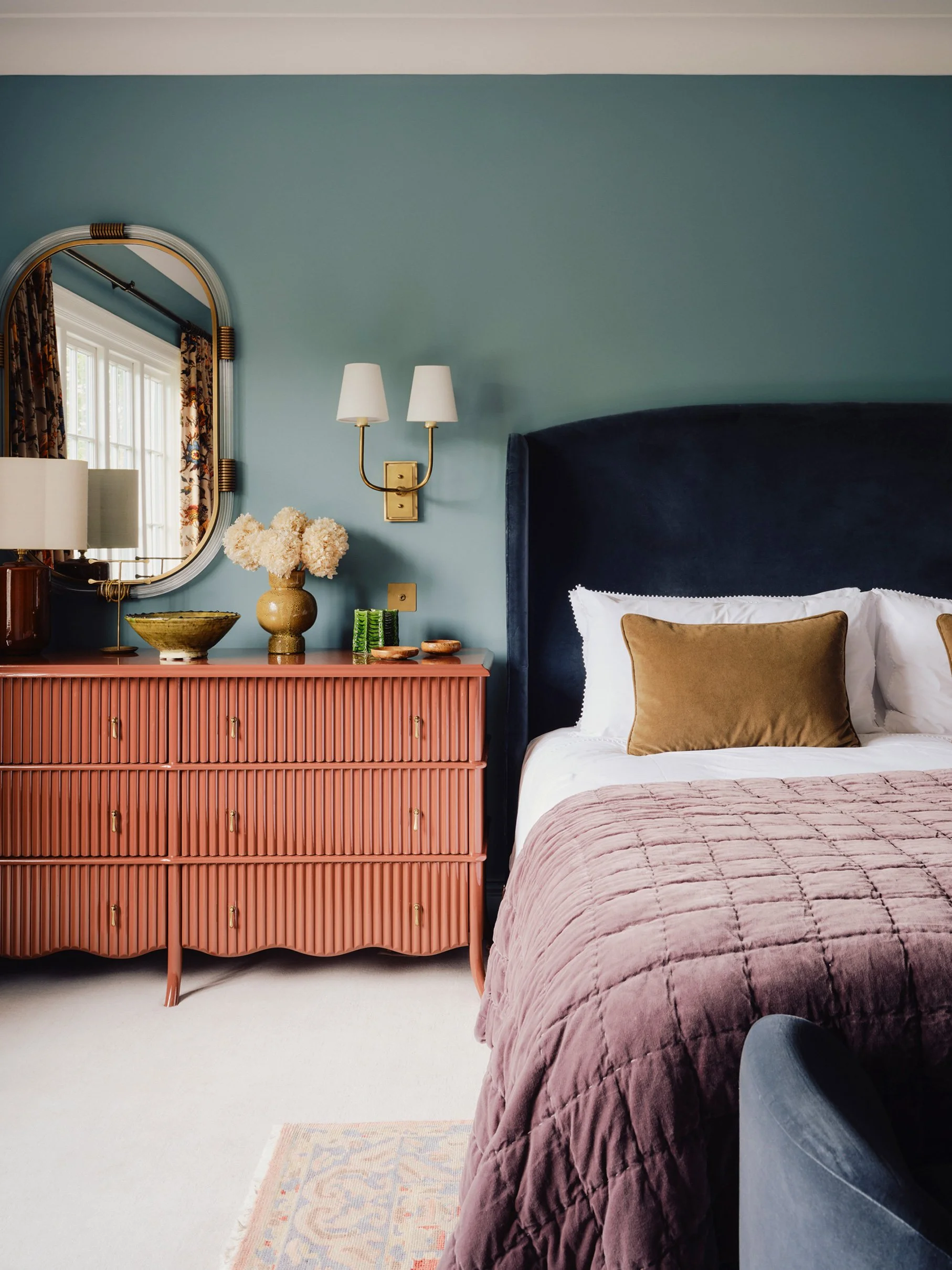 A bedroom designed by Imperfect Interiors with blue walls painted in Imperfect Interiors x Pilgrim Herring paint, a dark blue velvet headboard,  a pink velvet bedspread, a coral-coloured dresser with decorative items, a mirror,  with floral curtains
