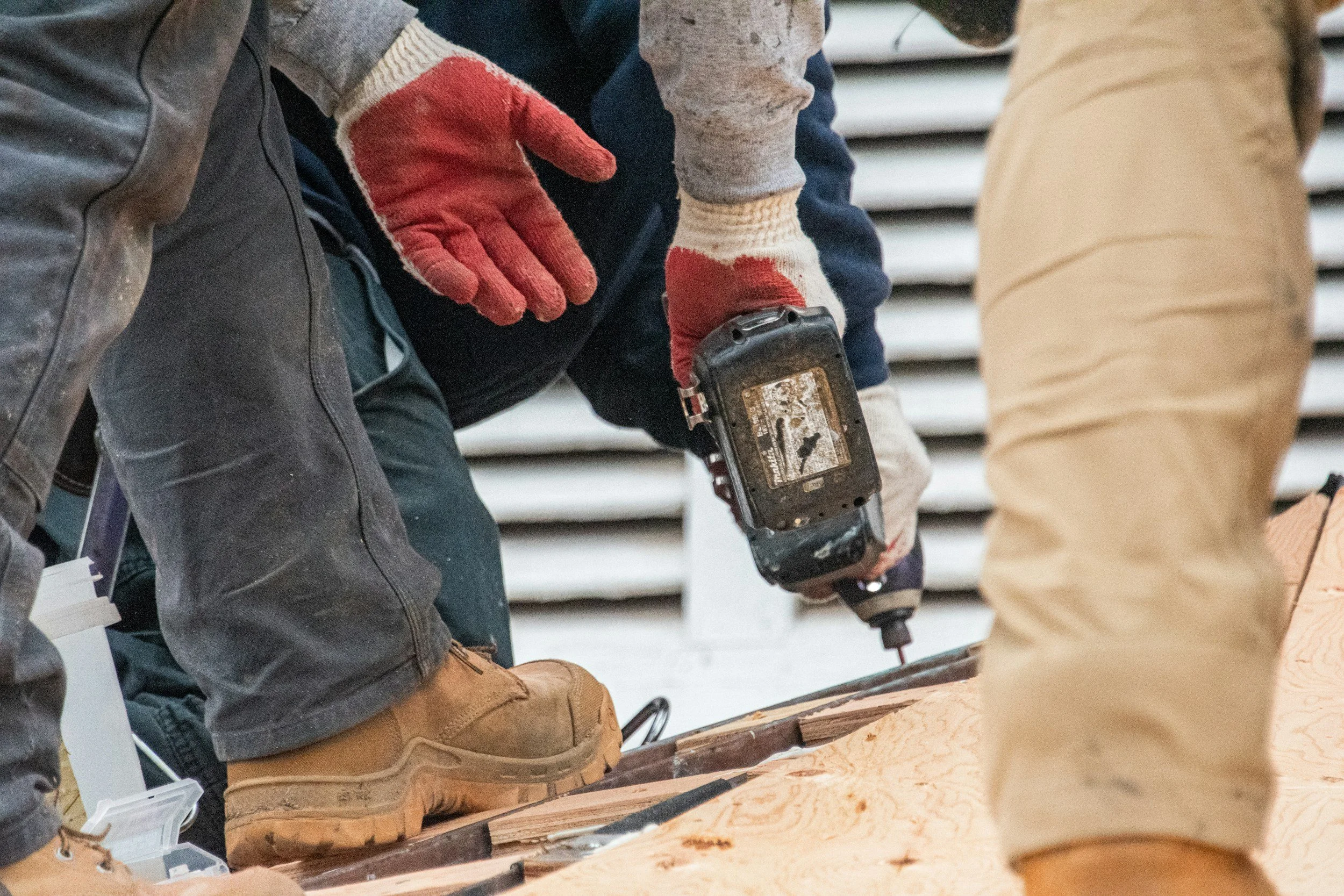Construction workers using a cordless drill to work on a wooden surface, dressed in work boots and gloves.