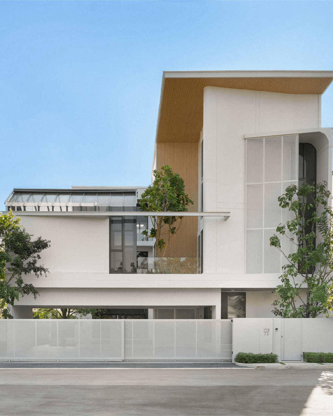 Modern multi-story residential building with white exterior walls, large windows, and green trees in front. Clear blue sky overhead.