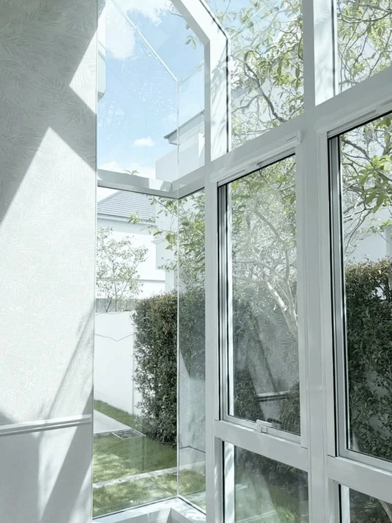 Bright, modern room with large glass windows and a glass door, showing a view of a garden with trees and bushes outside.